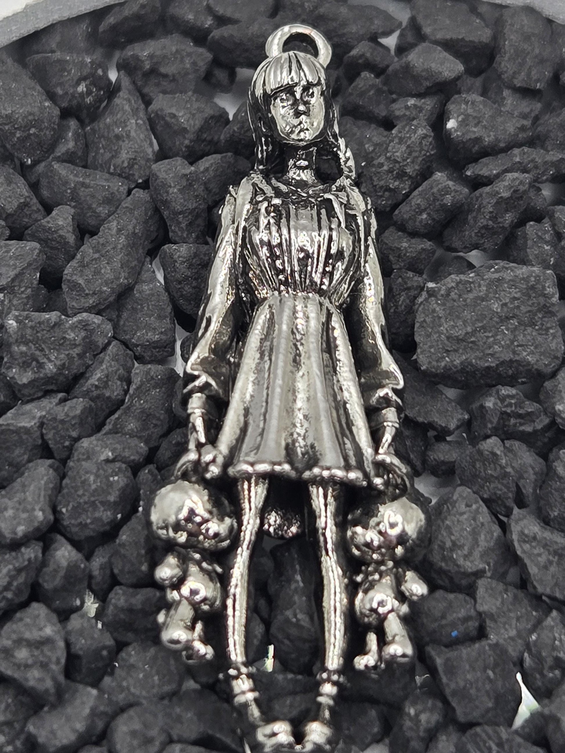 A silver-colored metal charm in the shape of a woman, with a long dress and a headpiece, is resting on a bed of small rocks.
