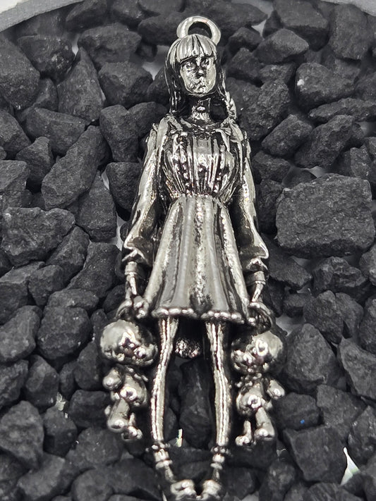 A silver-colored metal charm in the shape of a woman, with a long dress and a headpiece, is resting on a bed of small rocks.