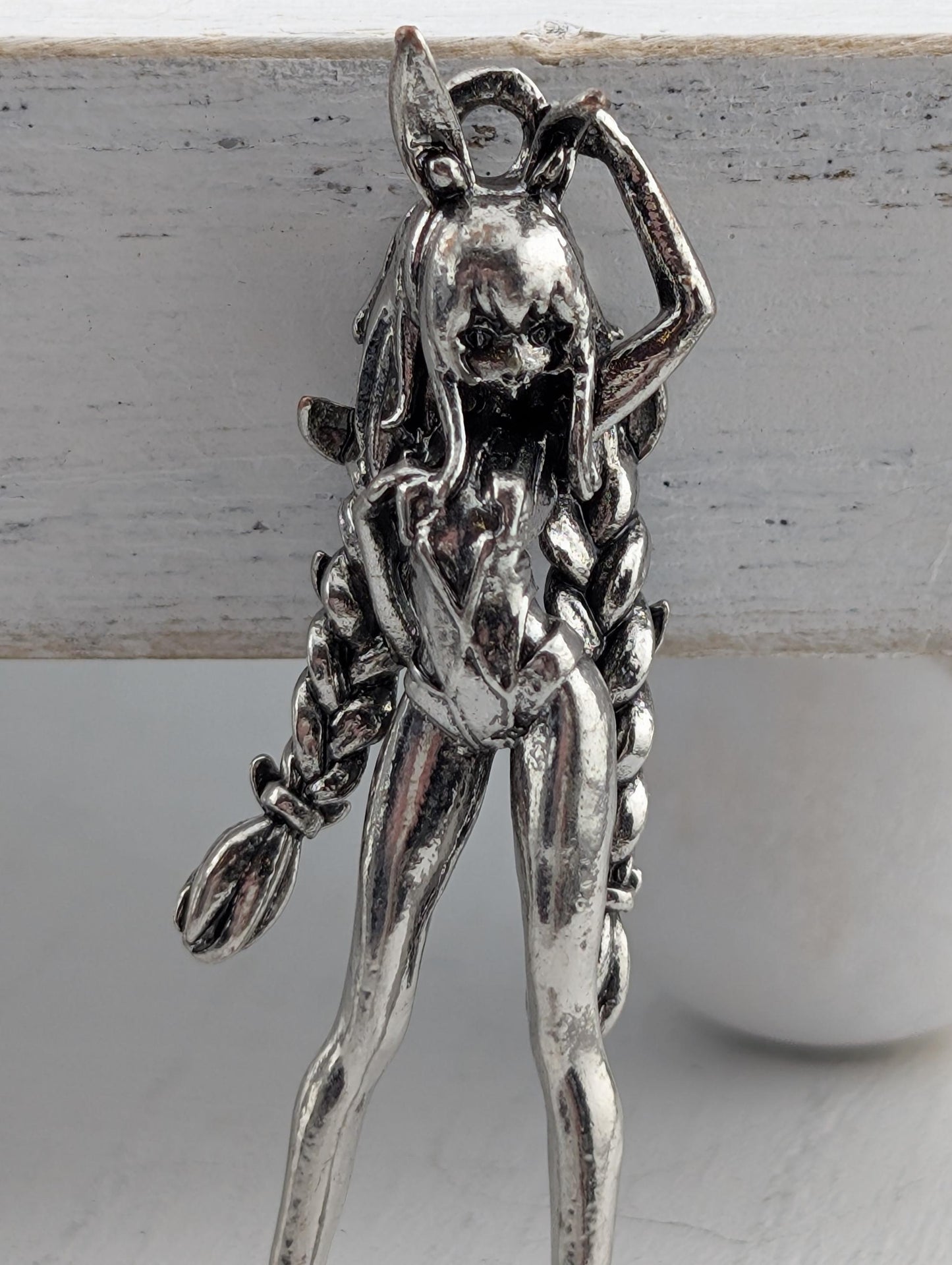 A silver-colored metal figurine of a woman with long braided hair, wearing a skirt and holding a broom.