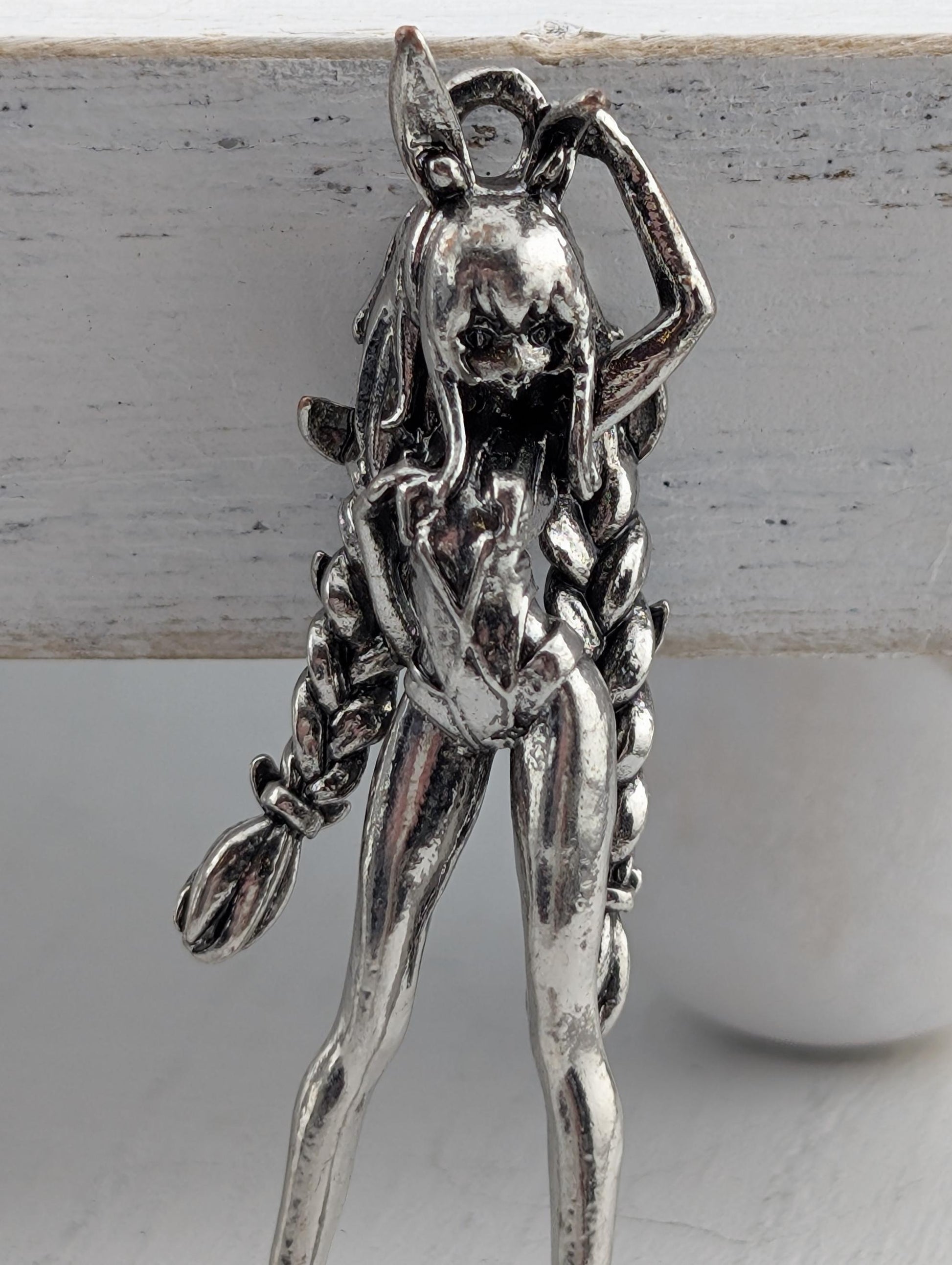 A silver-colored metal figurine of a woman with long braided hair, wearing a skirt and holding a broom.
