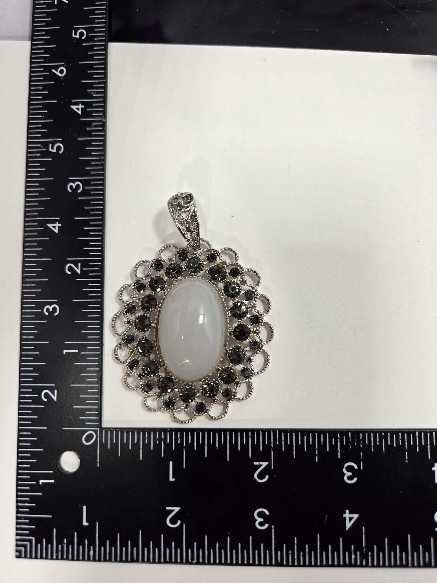 a pendant with a white stone and a decorative metal frame, placed next to a ruler for size comparison.
