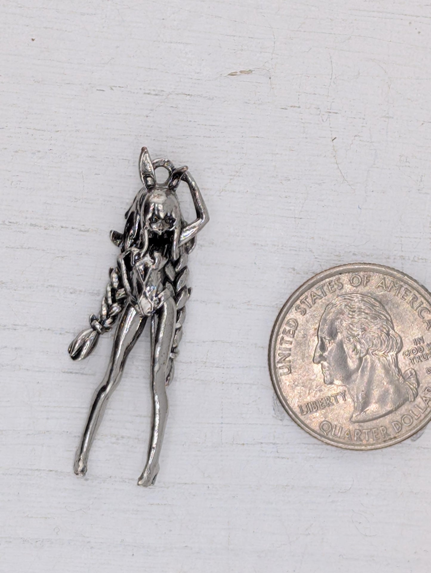 A silver-colored metal charm in the shape of a female figure is placed next to a United States quarter coin on a white surface.