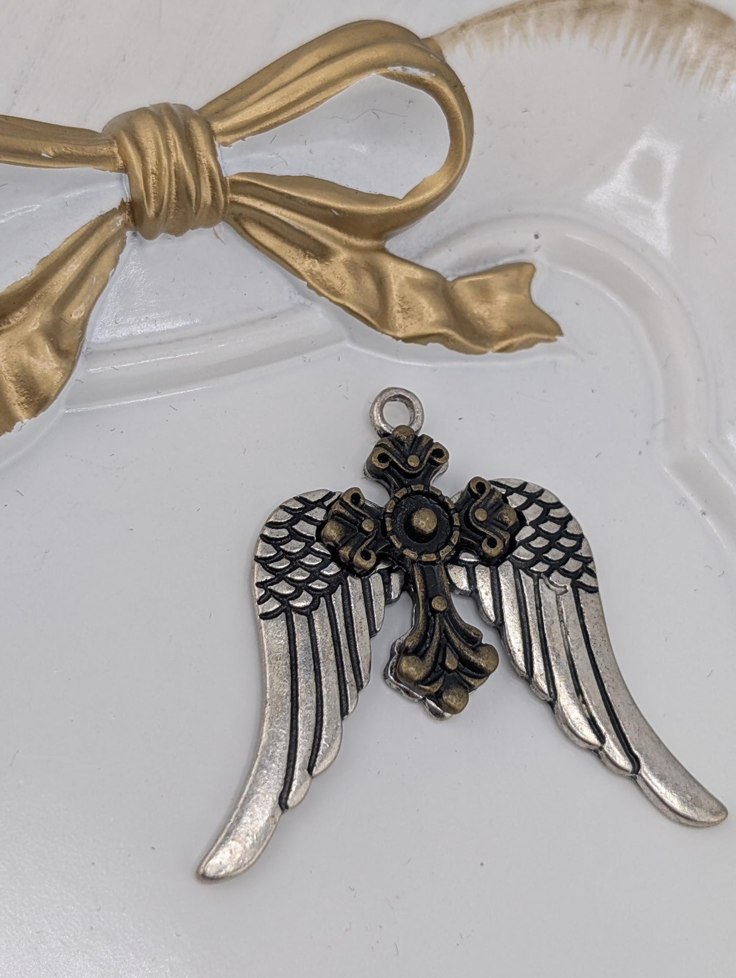 A decorative metal charm in the shape of a cross with wings, placed on a white surface next to a gold ribbon bow.