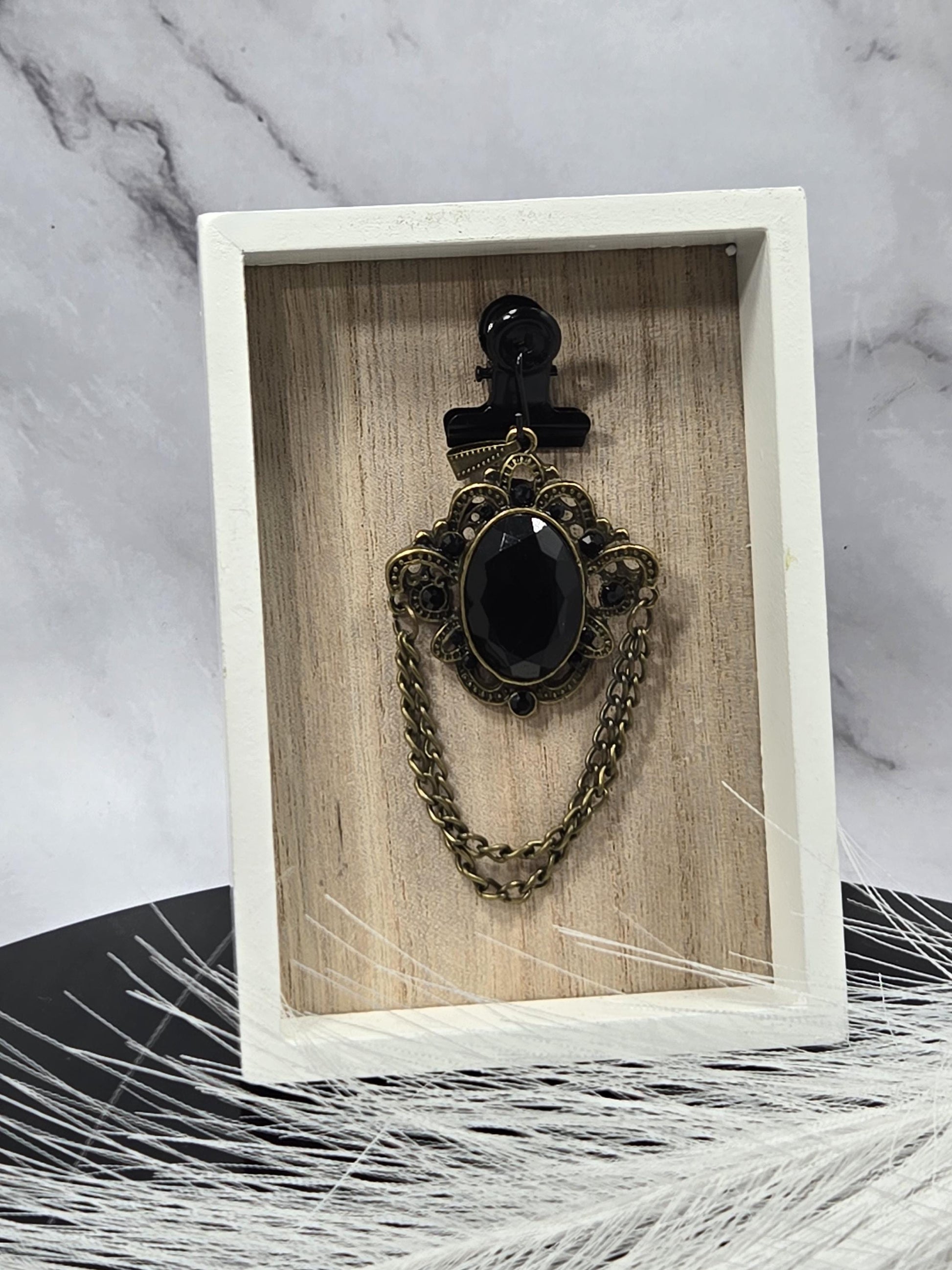 A white wooden box containing a black ornate piece of jewelry, possibly a necklace or bracelet, with a chain and a large black gemstone.