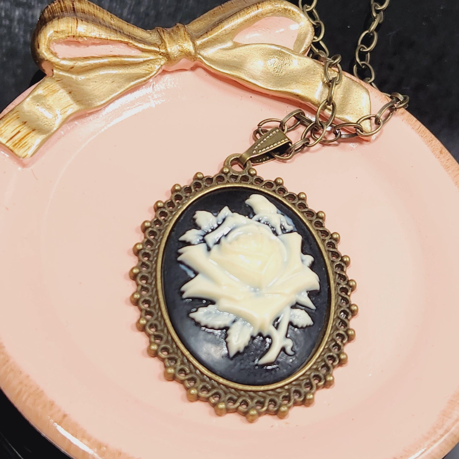 A necklace with a pendant featuring a white flower design is displayed on a pink plate, accompanied by a gold bow.