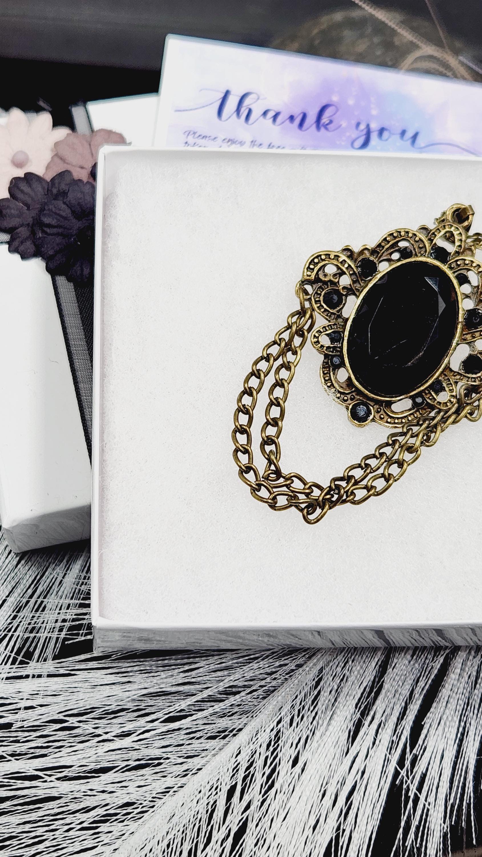 A close-up view of a decorative black and gold brooch with a chain, placed on a white surface.