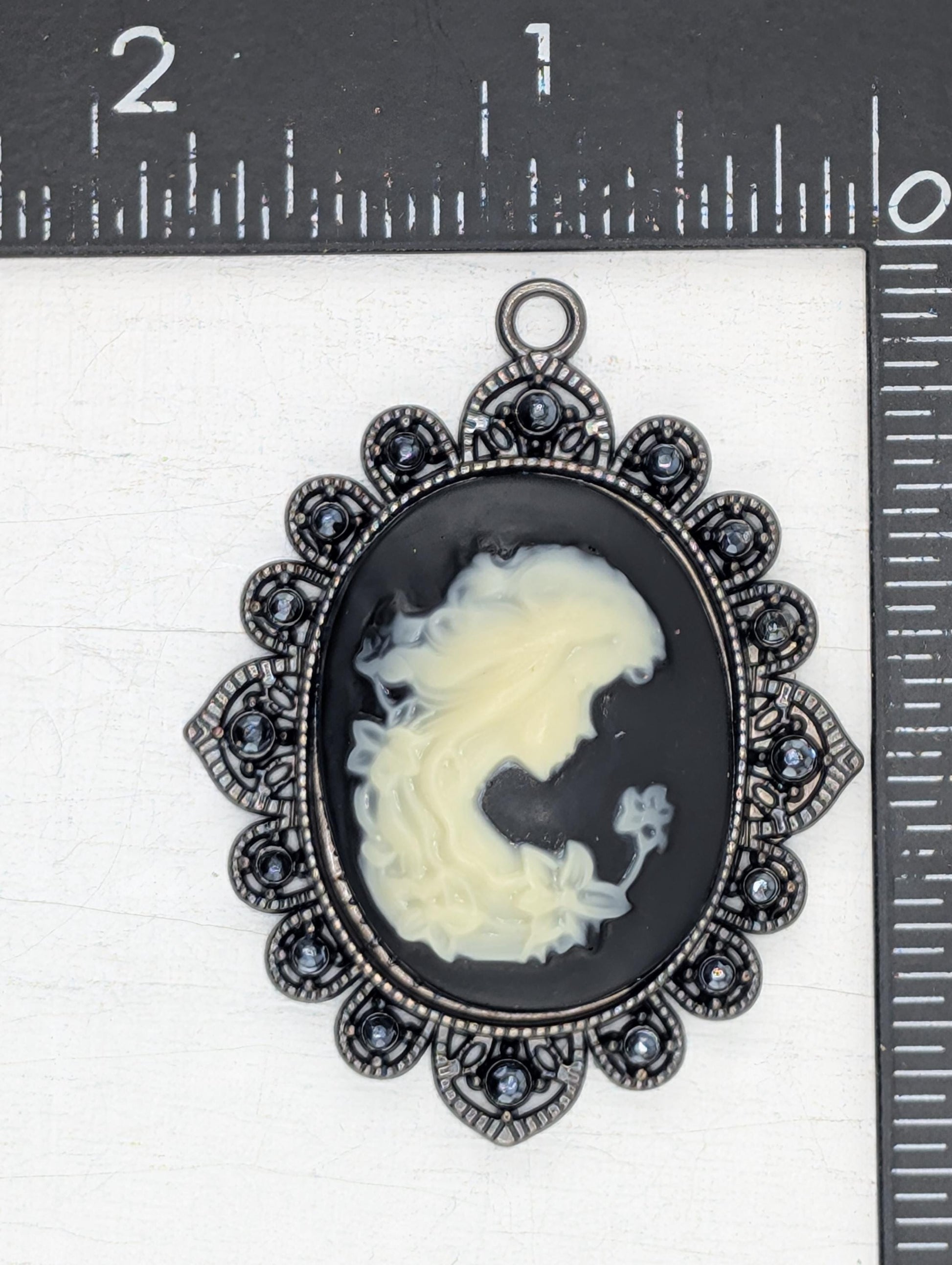 a pendant with a black oval background and a white silhouette of a woman&#39;s face. The pendant is adorned with a decorative metal frame and has small blue stones or crystals embedded in it. The pendant is placed on a white surface, and there is a ruler visible in the background.