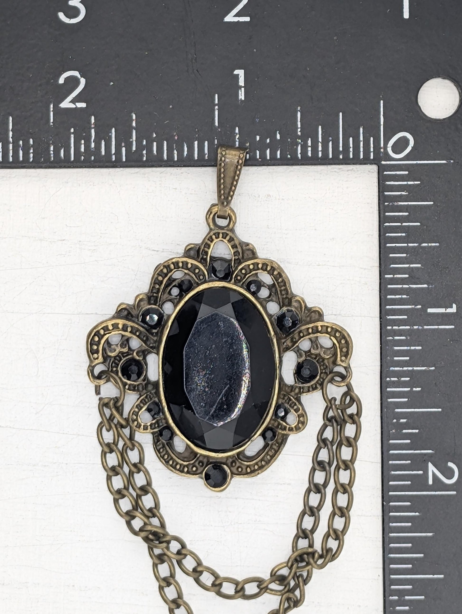 A black stone pendant with an ornate, decorative design and a chain is shown against a ruler for size comparison.