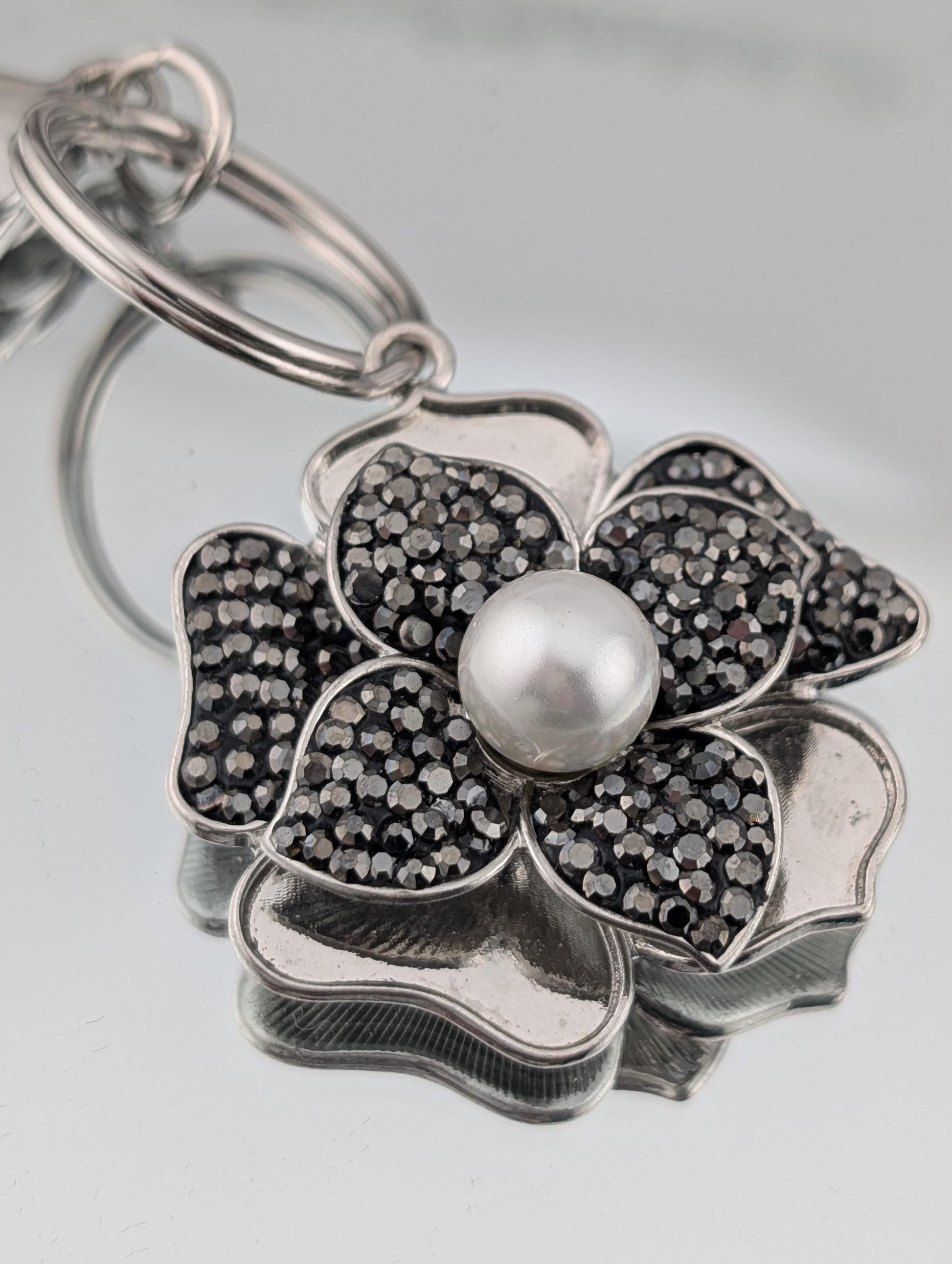 A silver-colored keychain with a flower-shaped pendant featuring a pearl at the center, surrounded by small black gemstones.