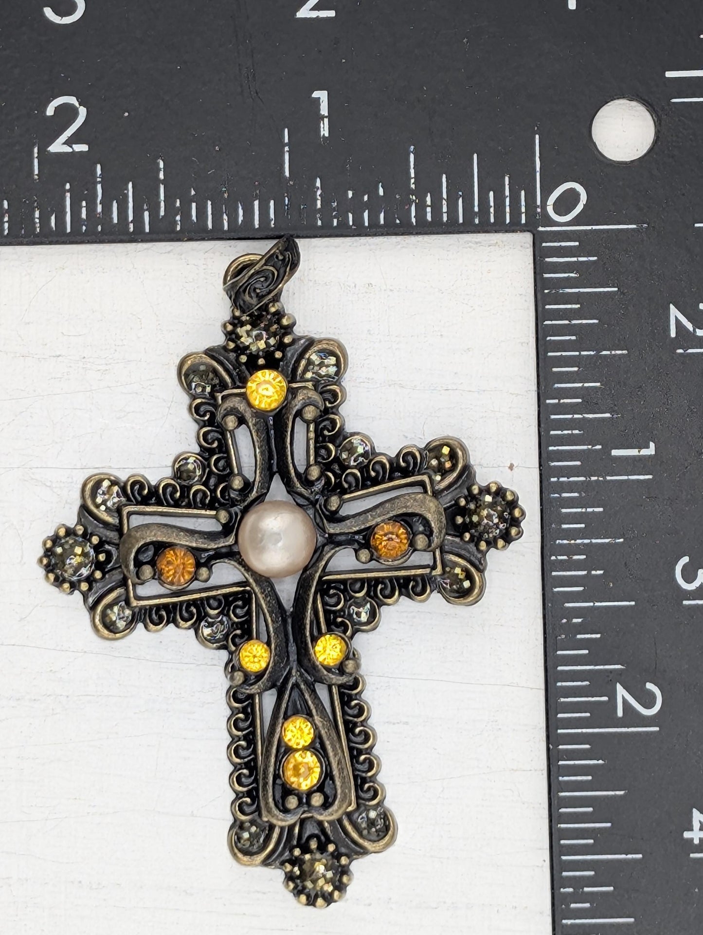 a decorative cross pendant with a pearl at its center, placed on a ruler for size comparison.