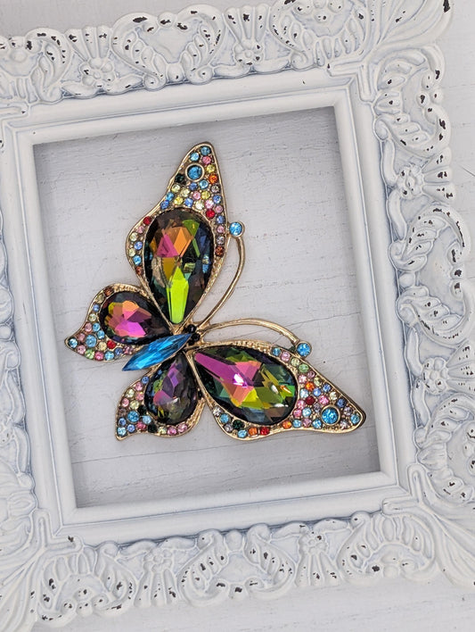 A colorful butterfly brooch with gemstones is displayed in a white ornate frame.