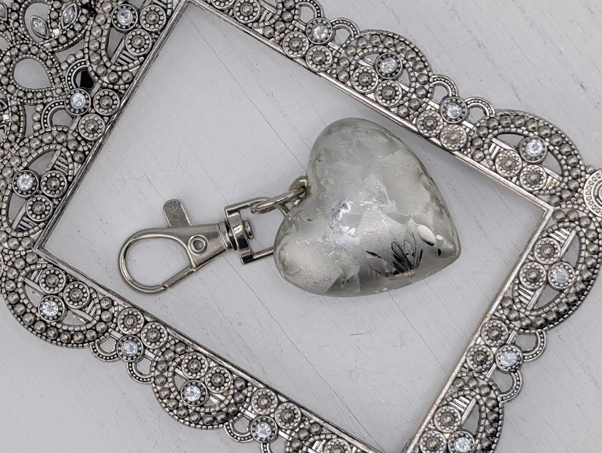 A silver heart-shaped charm with a silver clasp is displayed in a decorative silver frame with intricate designs.