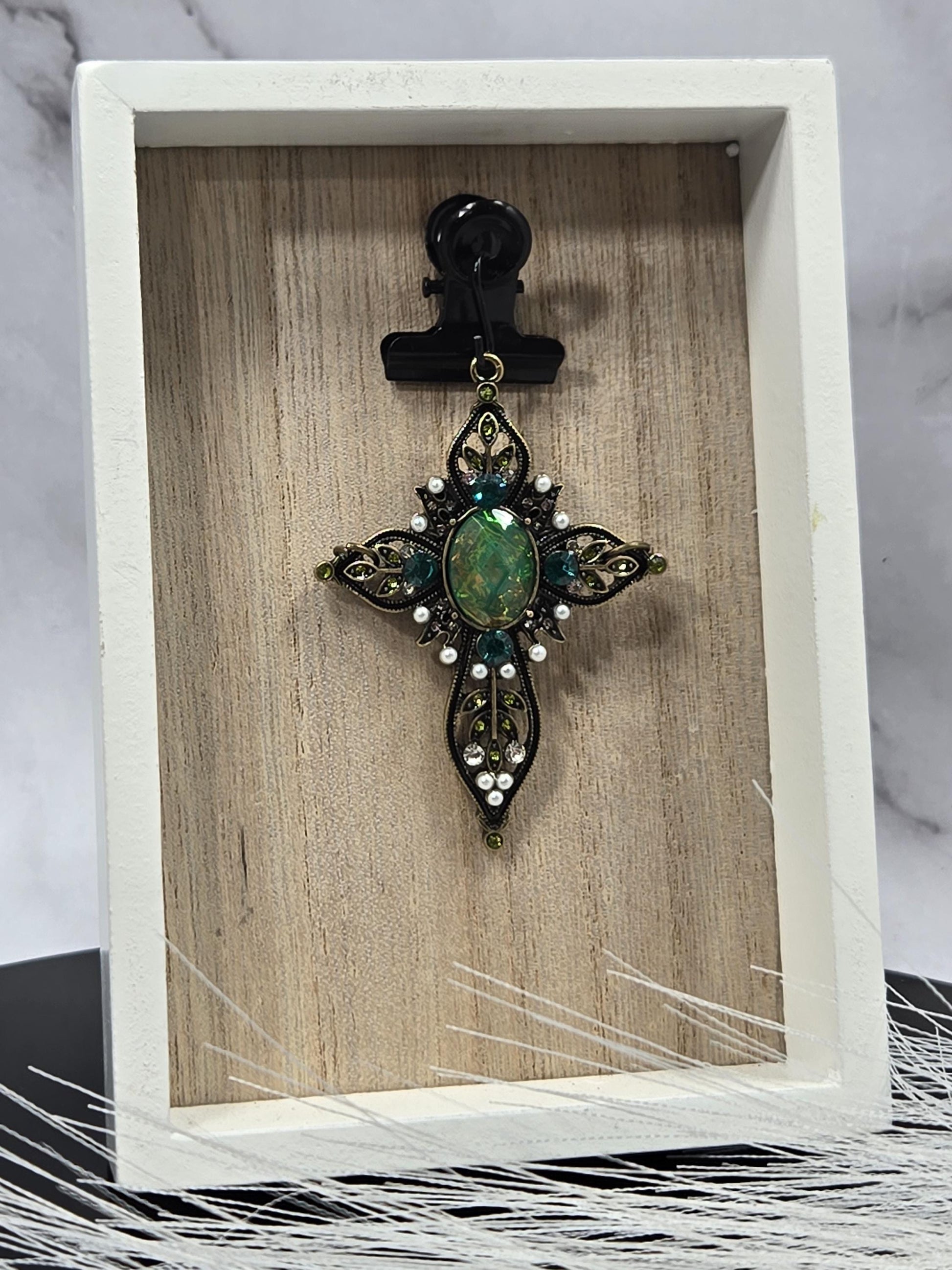 A decorative green gemstone brooch with intricate metalwork and white stones is displayed in a white wooden frame.