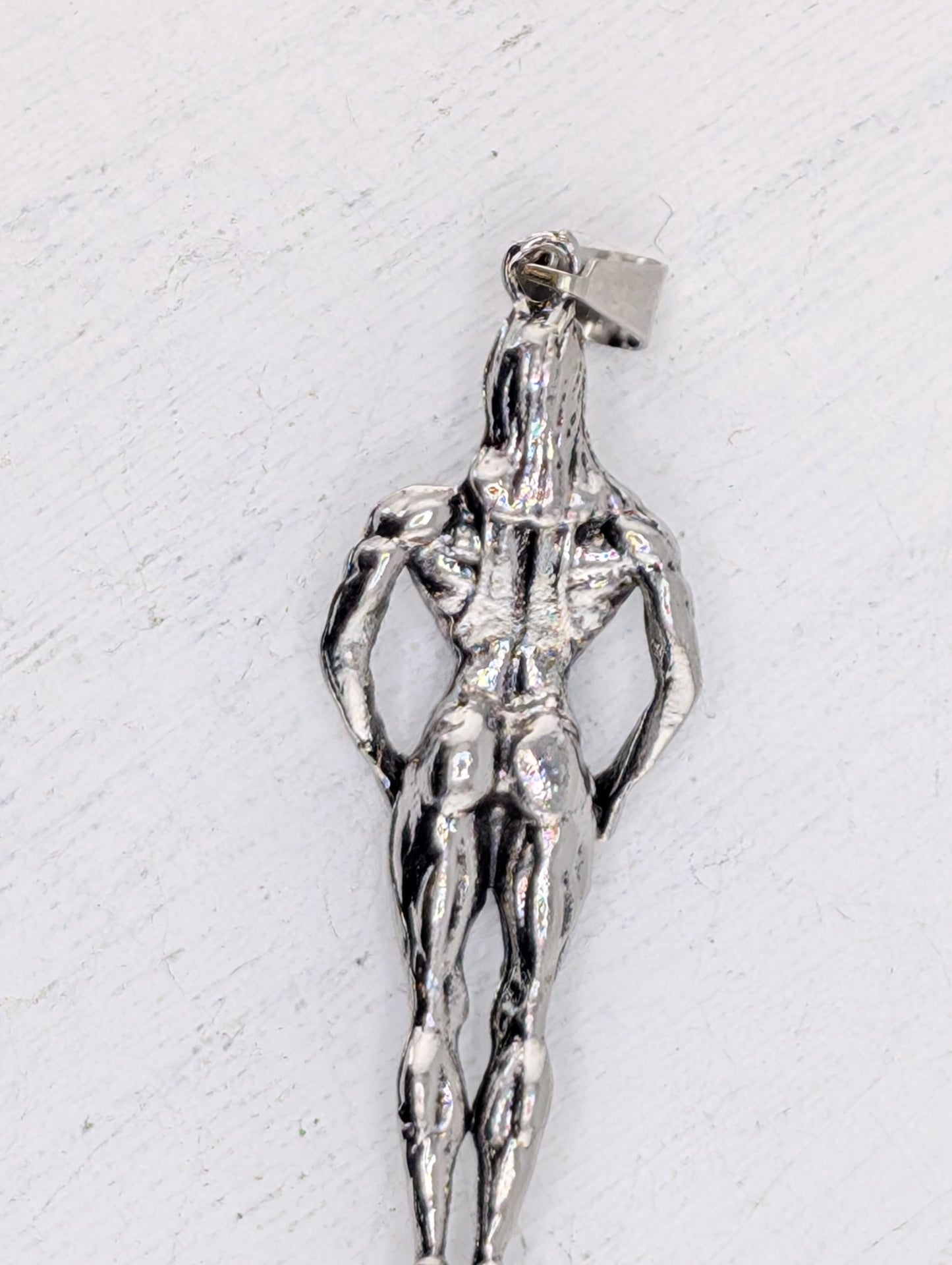 A silver-colored pendant in the shape of a muscular male figure, with a metallic sheen and a metallic clasp.