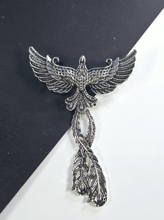 A silver-colored metal brooch with an intricate design, featuring a bird-like figure with outstretched wings and decorative feathers.