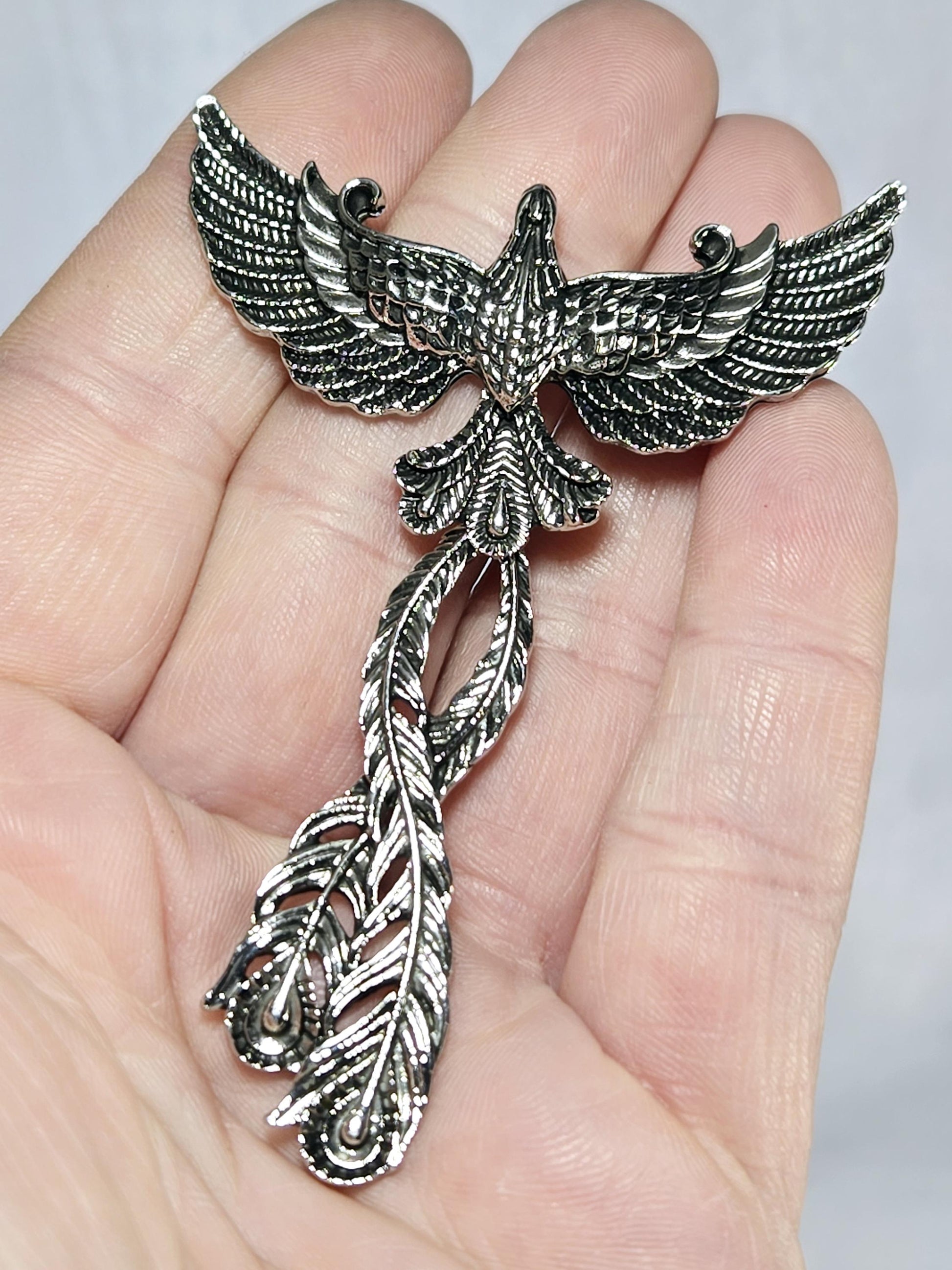 A hand holding a silver-colored metal pin or brooch in the shape of a bird with wings spread out.