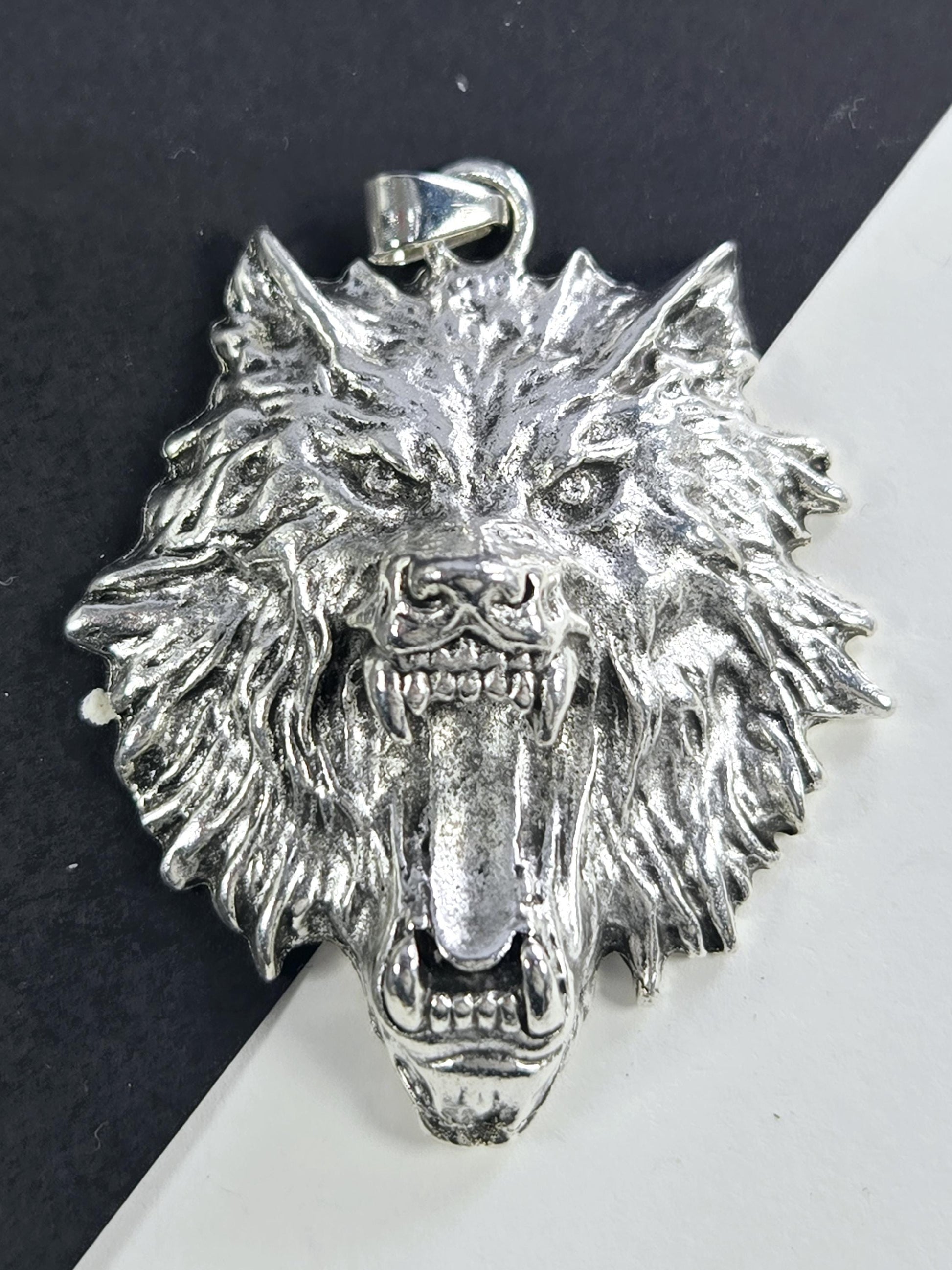 A silver-colored metal pendant in the shape of a fierce-looking animal head, possibly a wolf or a lion, with sharp teeth and an intense expression.