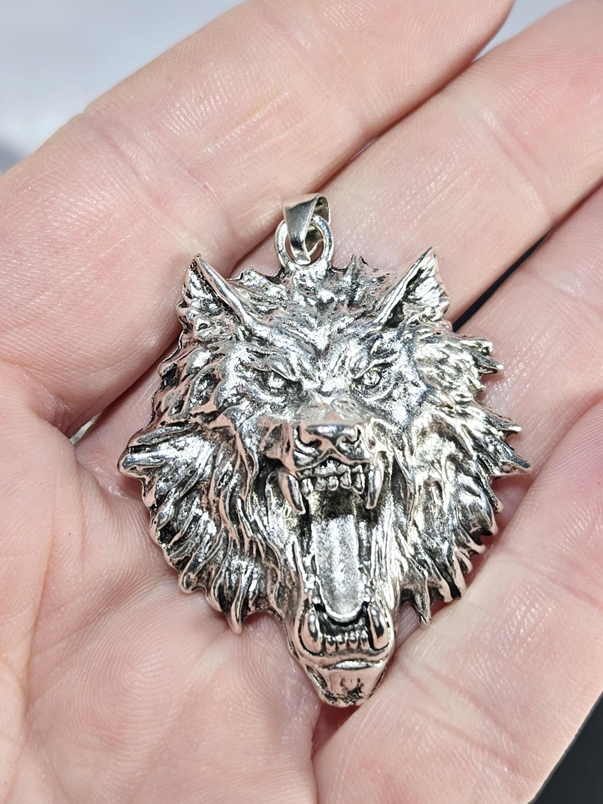 A close-up image of a person&#39;s hand holding a large, detailed silver pendant in the shape of a fierce-looking wolf&#39;s head.