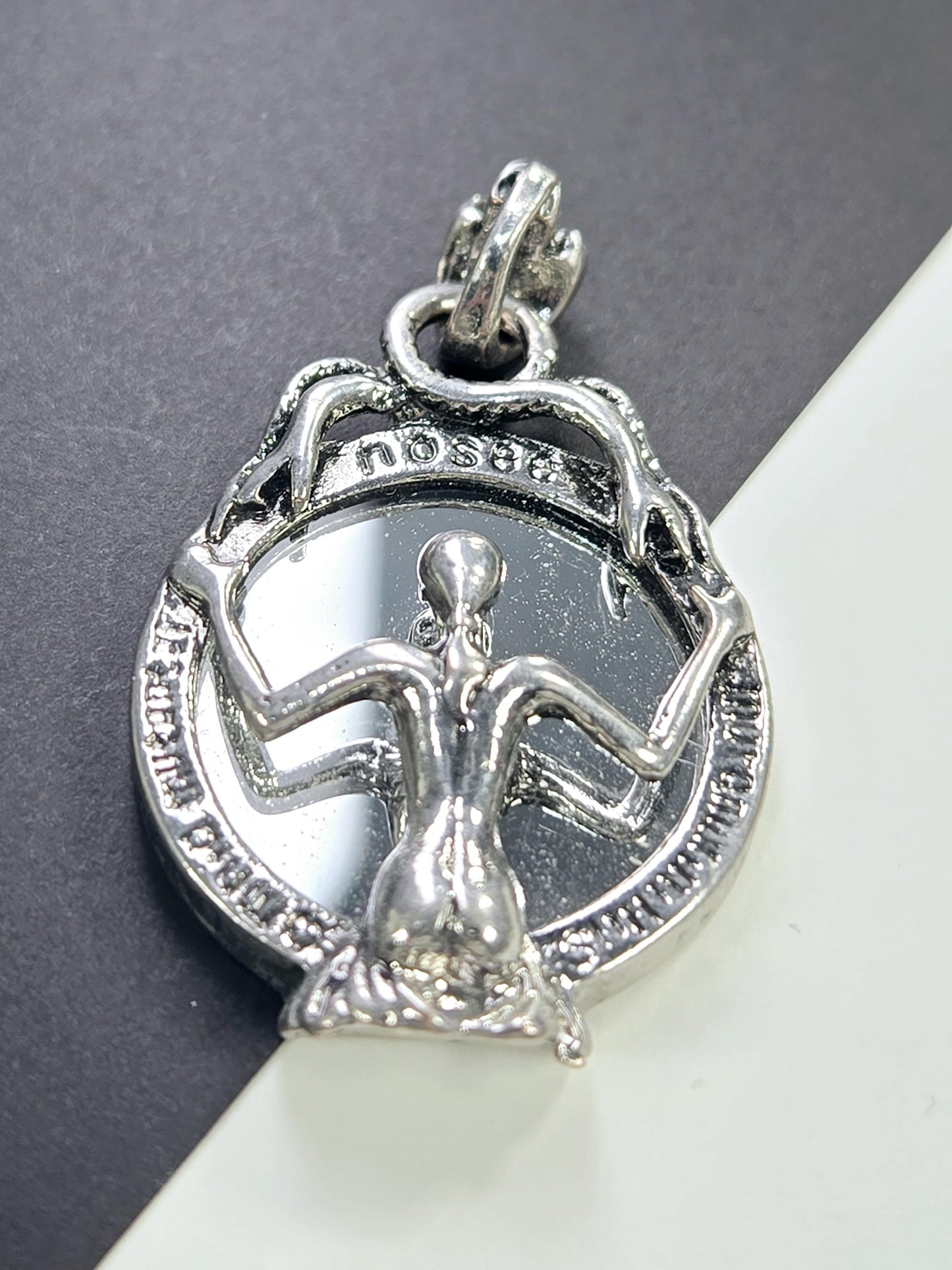 A silver-colored pendant or charm with an intricate design, featuring a figure or symbol within a circular frame.