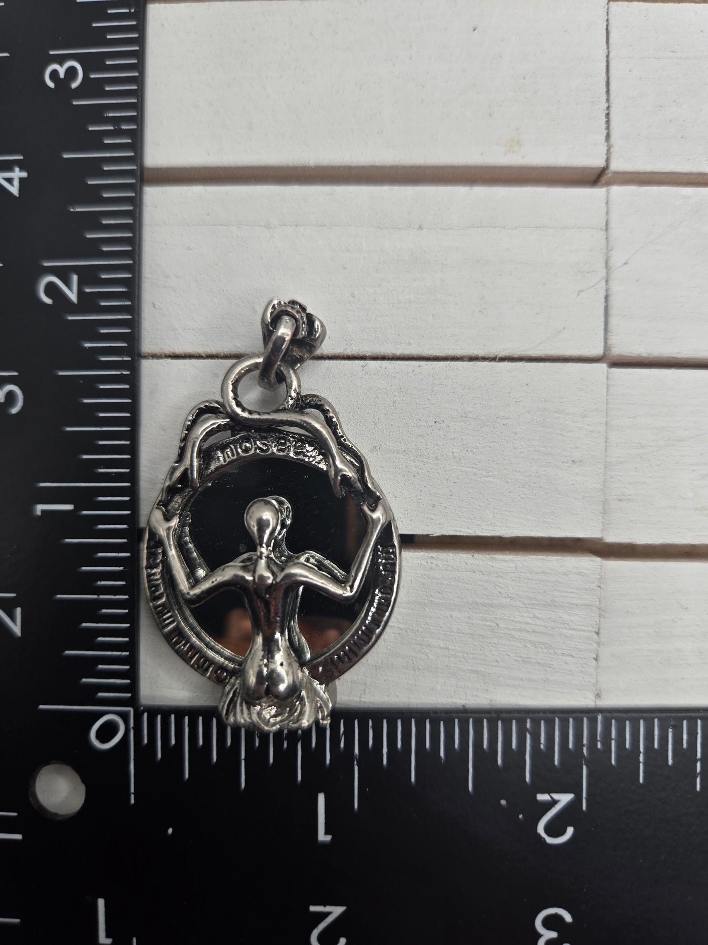 A silver-colored metal charm or pendant is placed next to a ruler on a white surface.