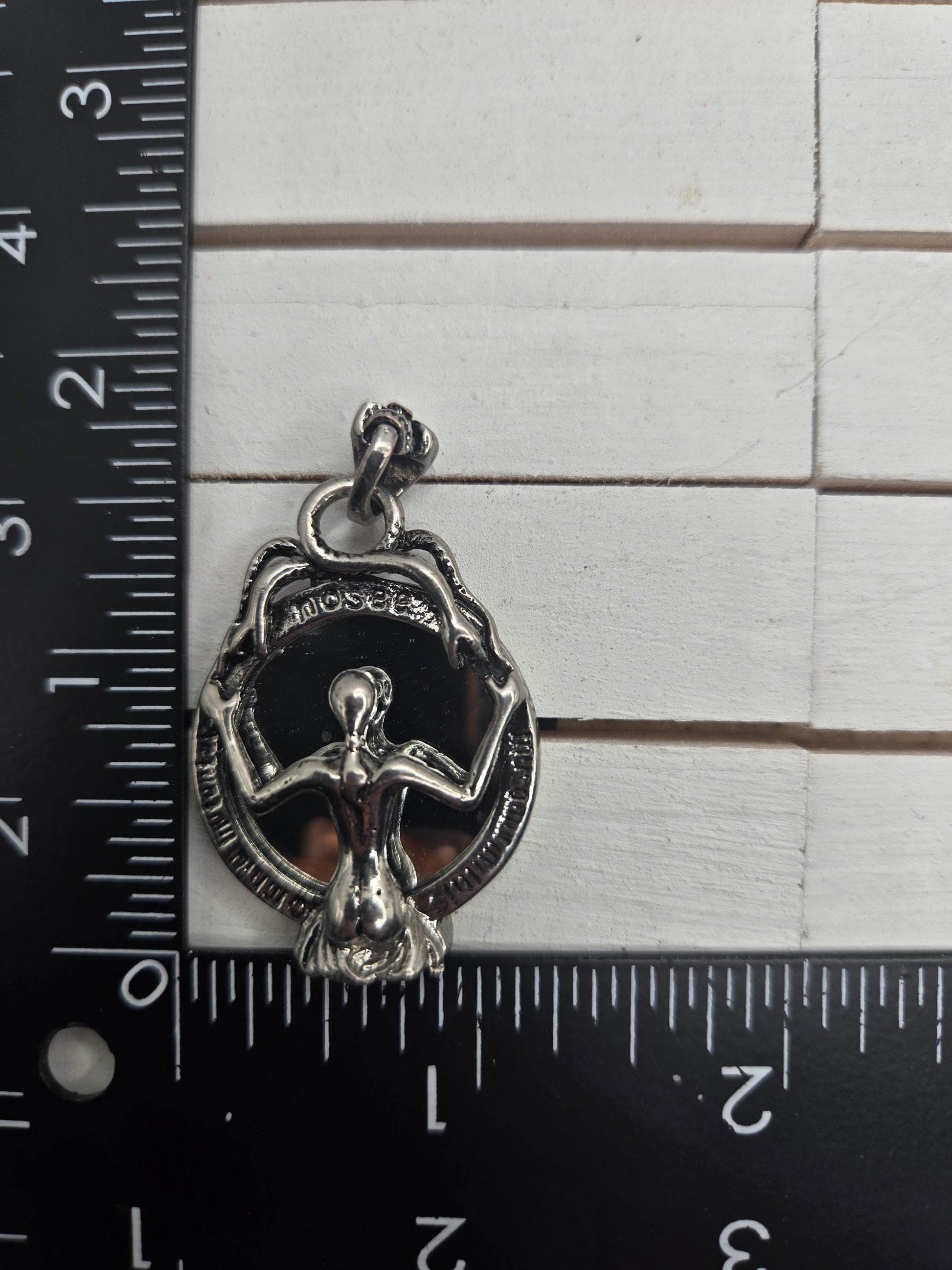 A silver-colored metal charm or pendant is placed next to a ruler on a white surface.