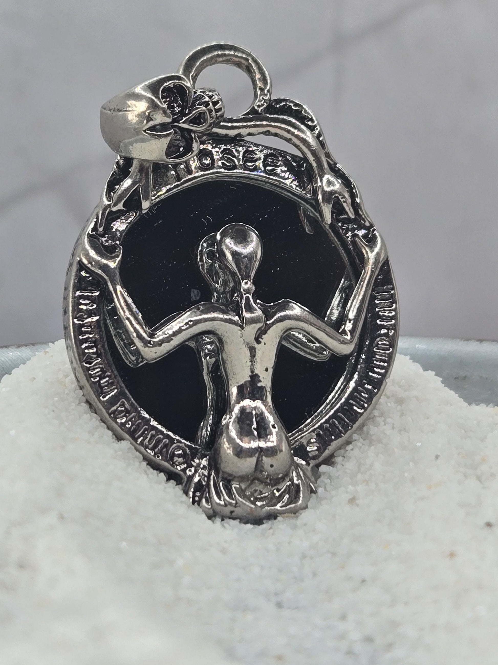 A silver-colored pendant or charm in the shape of a skull, with a black background and a decorative design around it.