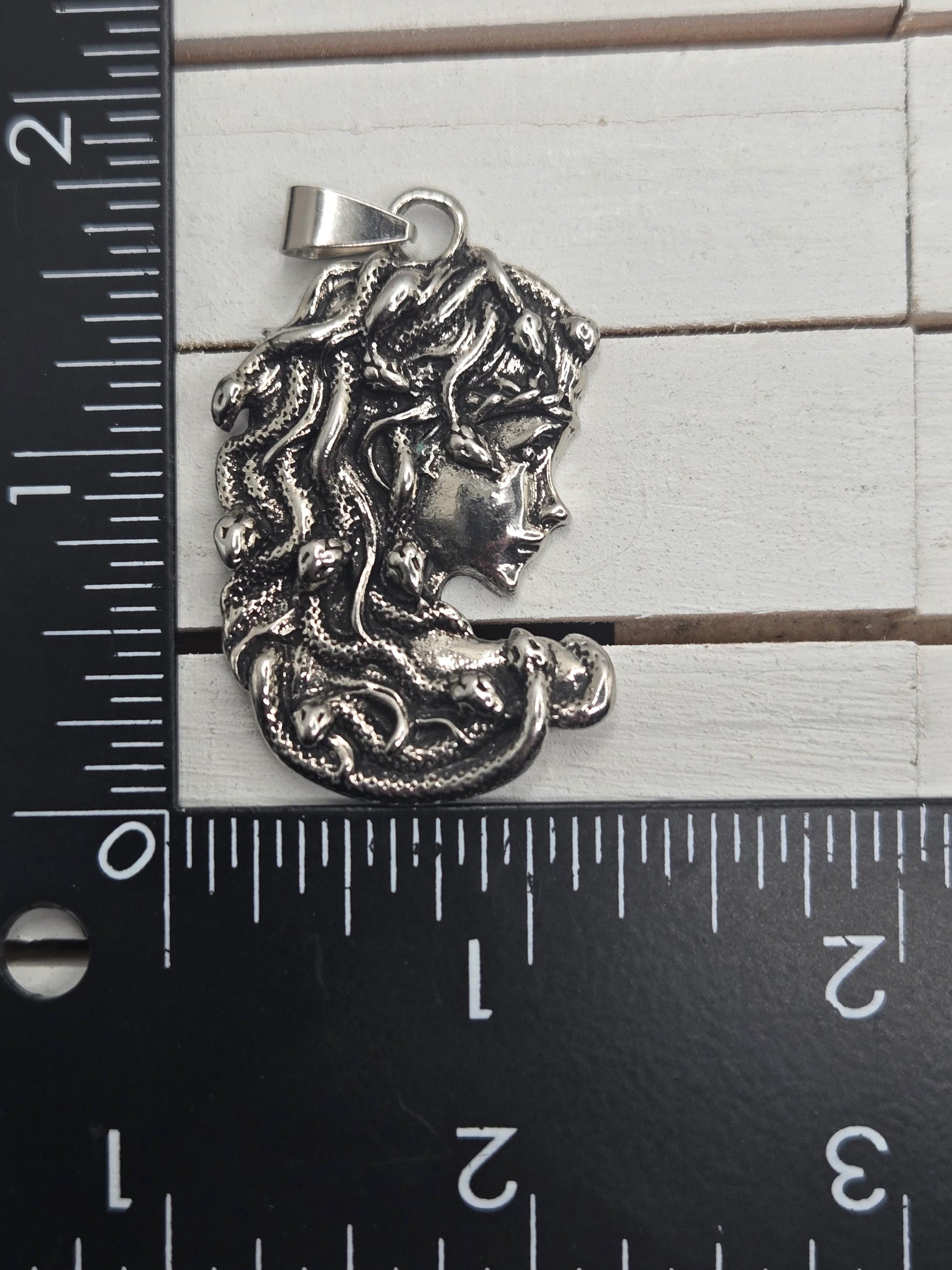 A silver-colored pendant in the shape of a woman&#39;s head with snakes wrapped around it, placed next to a ruler for size comparison.