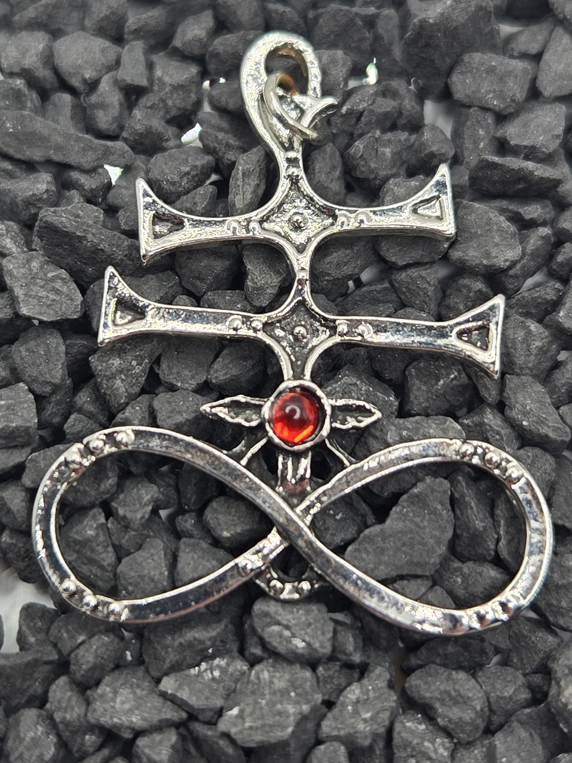 A silver pendant with a red gemstone, featuring an intricate design, is resting on a bed of small rocks.