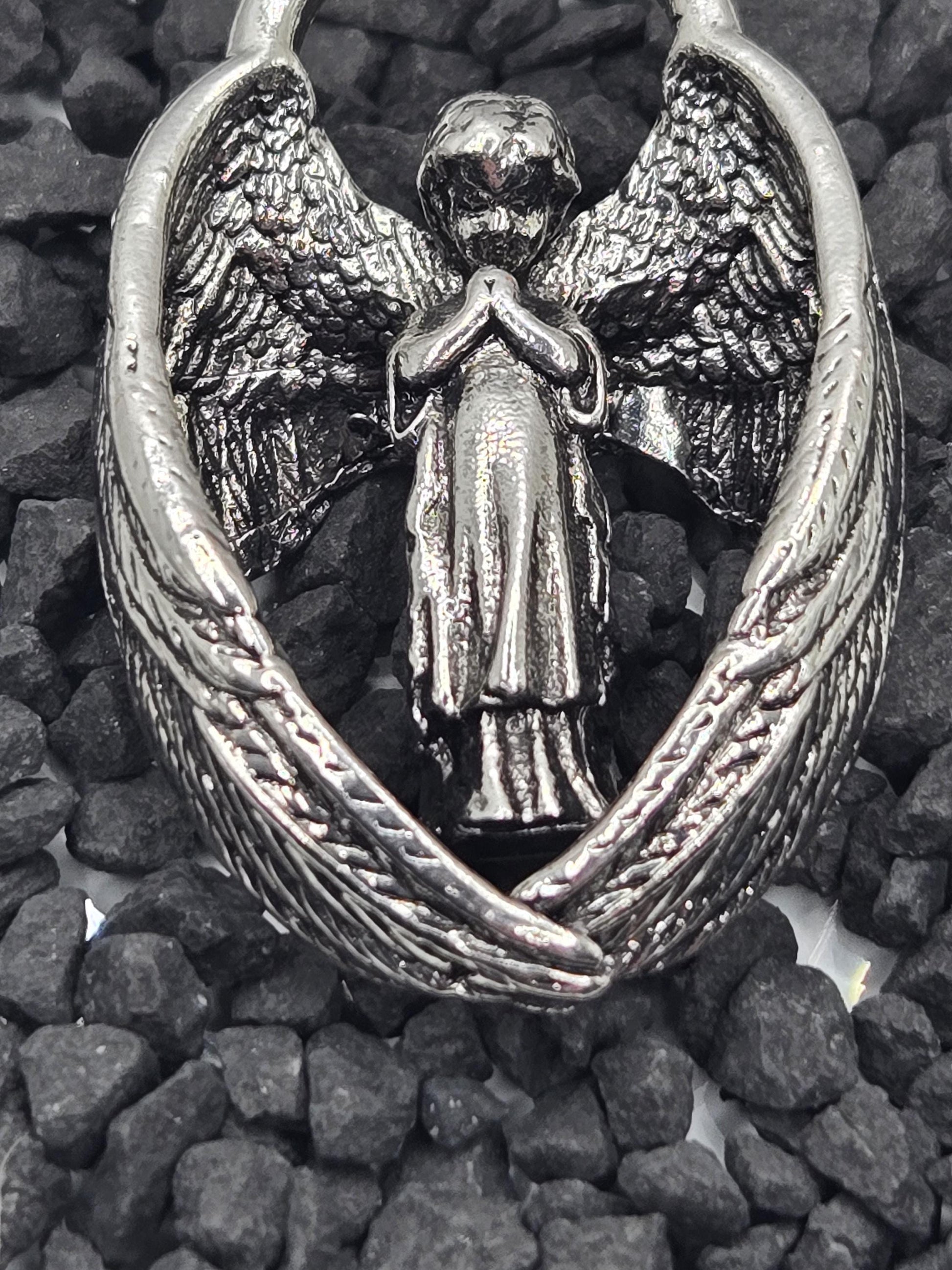 A silver-colored metal pendant in the shape of a winged figure, resting on a bed of small rocks.