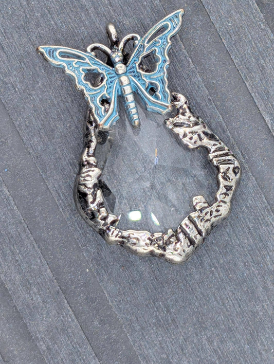 A silver butterfly-shaped pendant with a clear gemstone in the center, resting on a gray surface.