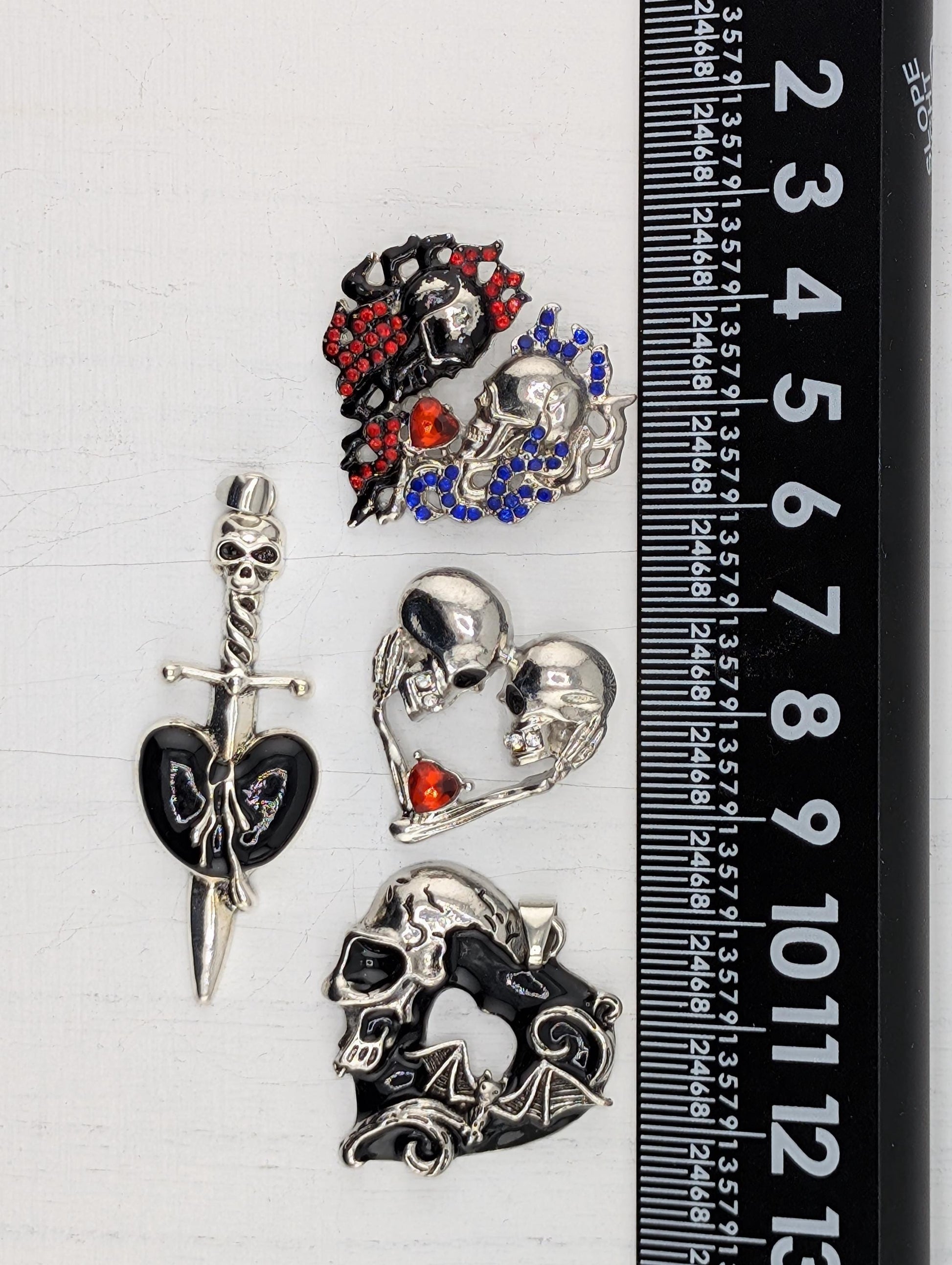 a collection of ornate metal pieces, including a skull-shaped brooch, a sword-shaped brooch, and a skull-shaped pendant, all adorned with colorful gemstones. The pieces are displayed on a ruler for scale, indicating their size relative to each other.