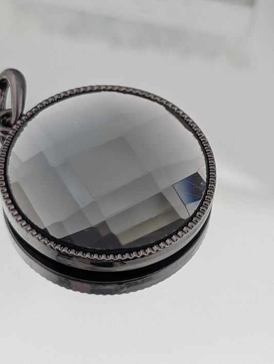 A close-up view of a round, black, and silver pendant with a clear, multicolored glass or crystal embedded in it.