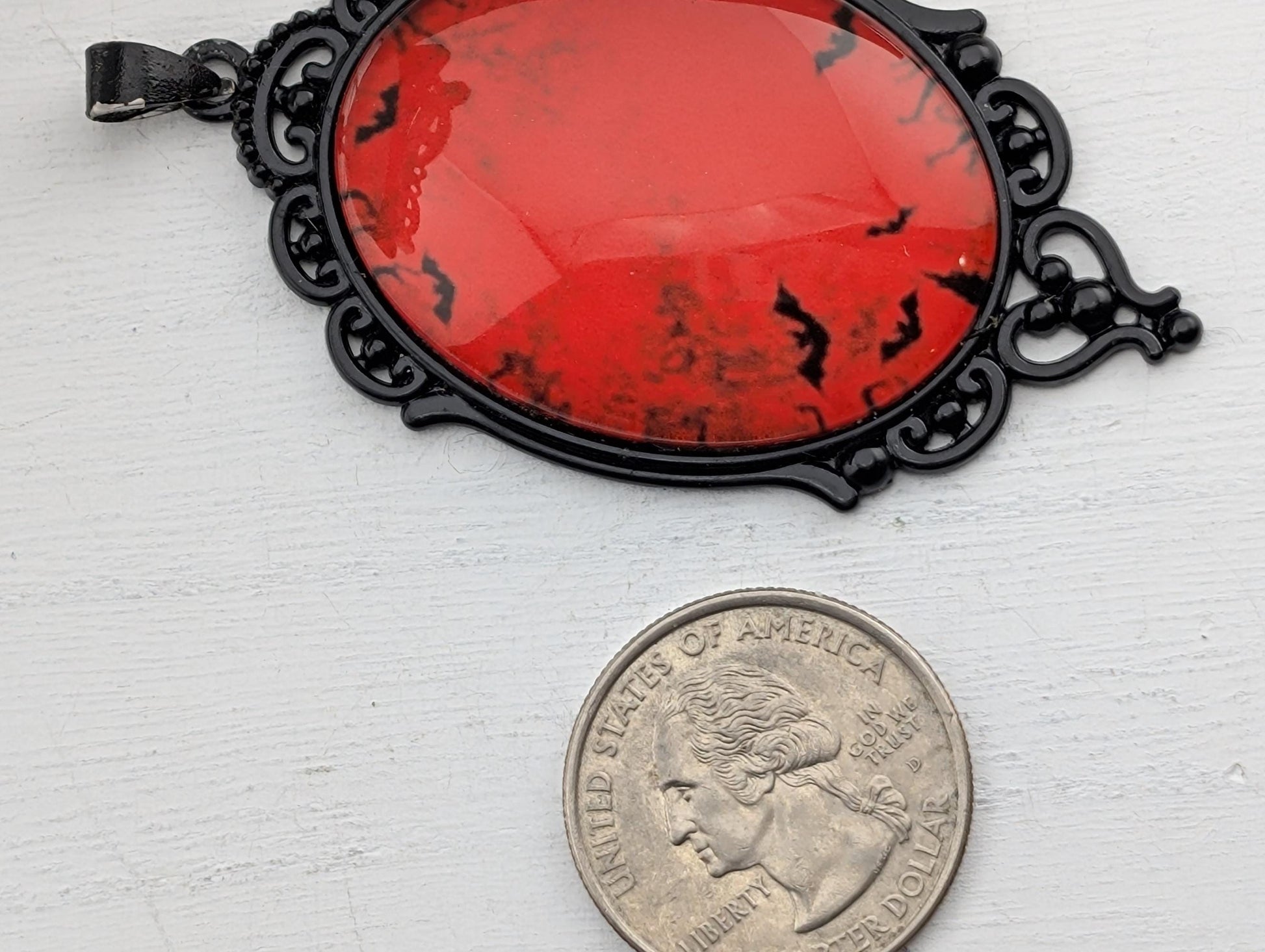 a large oval-shaped pendant with a red background and black bat-like designs, and a United States quarter is placed next to it on a white surface.
