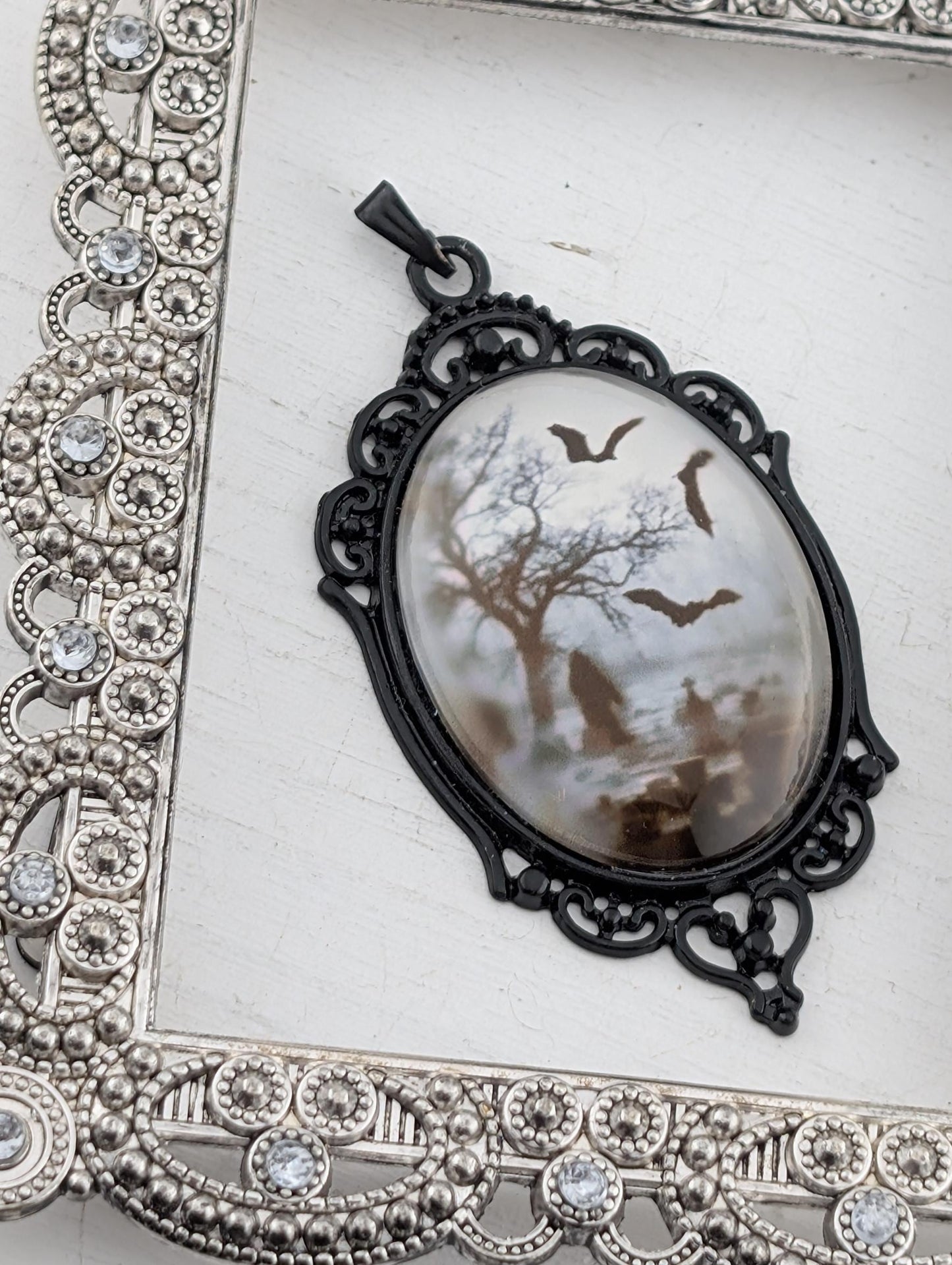 A pendant with a black metal frame and a white oval-shaped stone or glass piece featuring a bat silhouette and a snowy landscape.