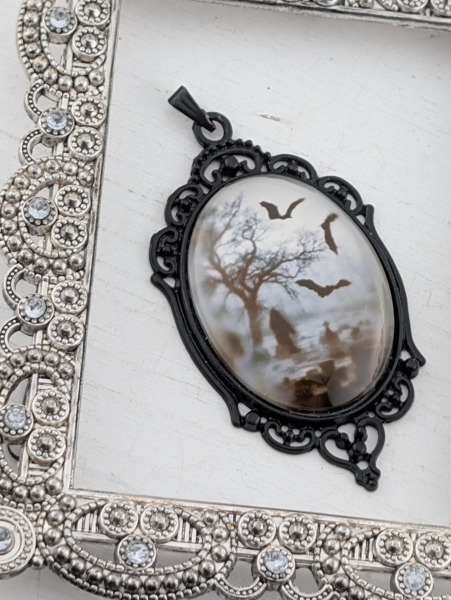 A pendant with a black metal frame and a white oval-shaped stone or glass piece featuring a bat silhouette and a snowy landscape.