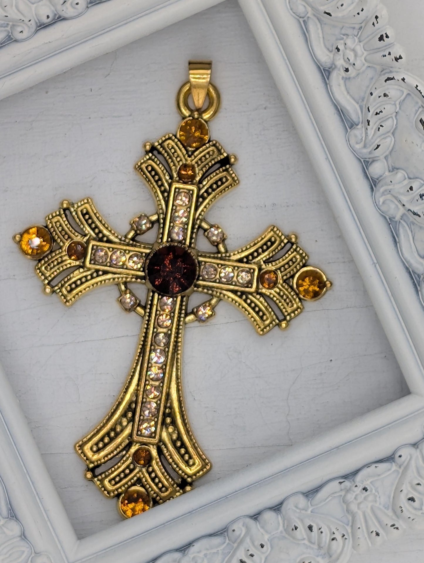 A decorative gold cross pendant with gemstones is displayed in a white ornate frame.