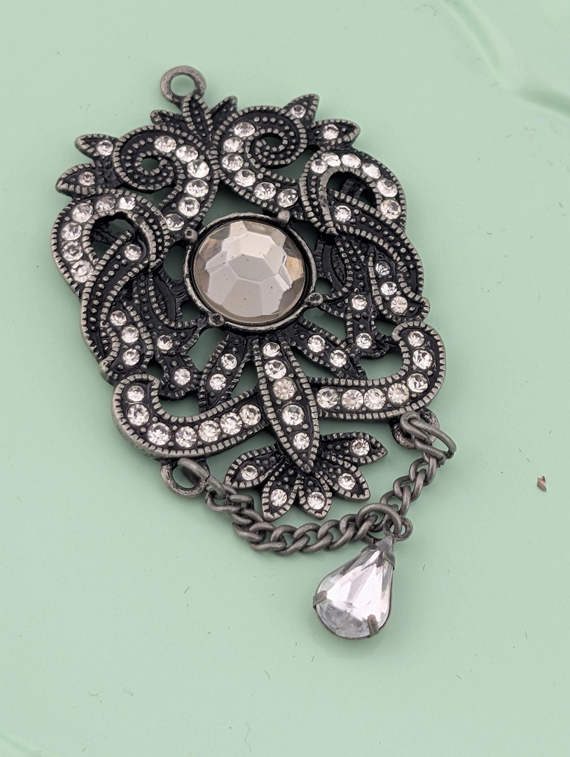 A decorative pendant with a large crystal at the center, surrounded by intricate floral and leaf designs, and a dangling crystal piece.