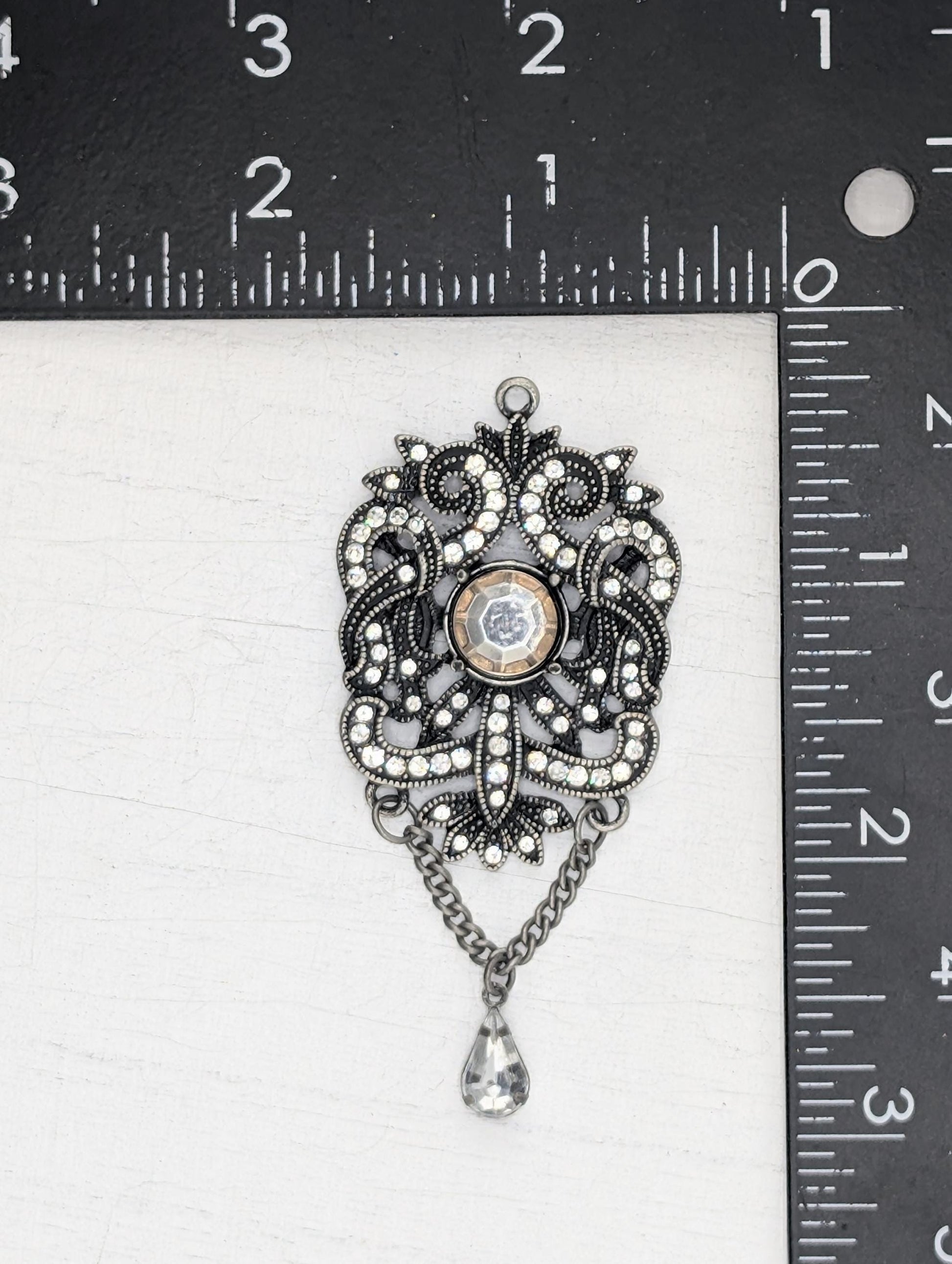 A decorative pendant with a floral design and a central gemstone is shown, placed above a ruler for scale.