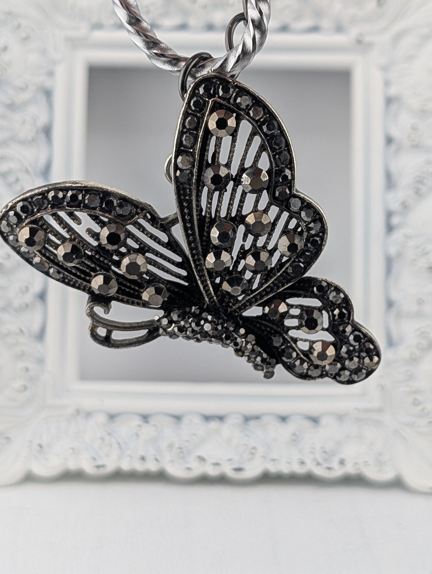 A decorative butterfly pendant with intricate black and silver designs, including rhinestones or crystals, hanging from a chain.