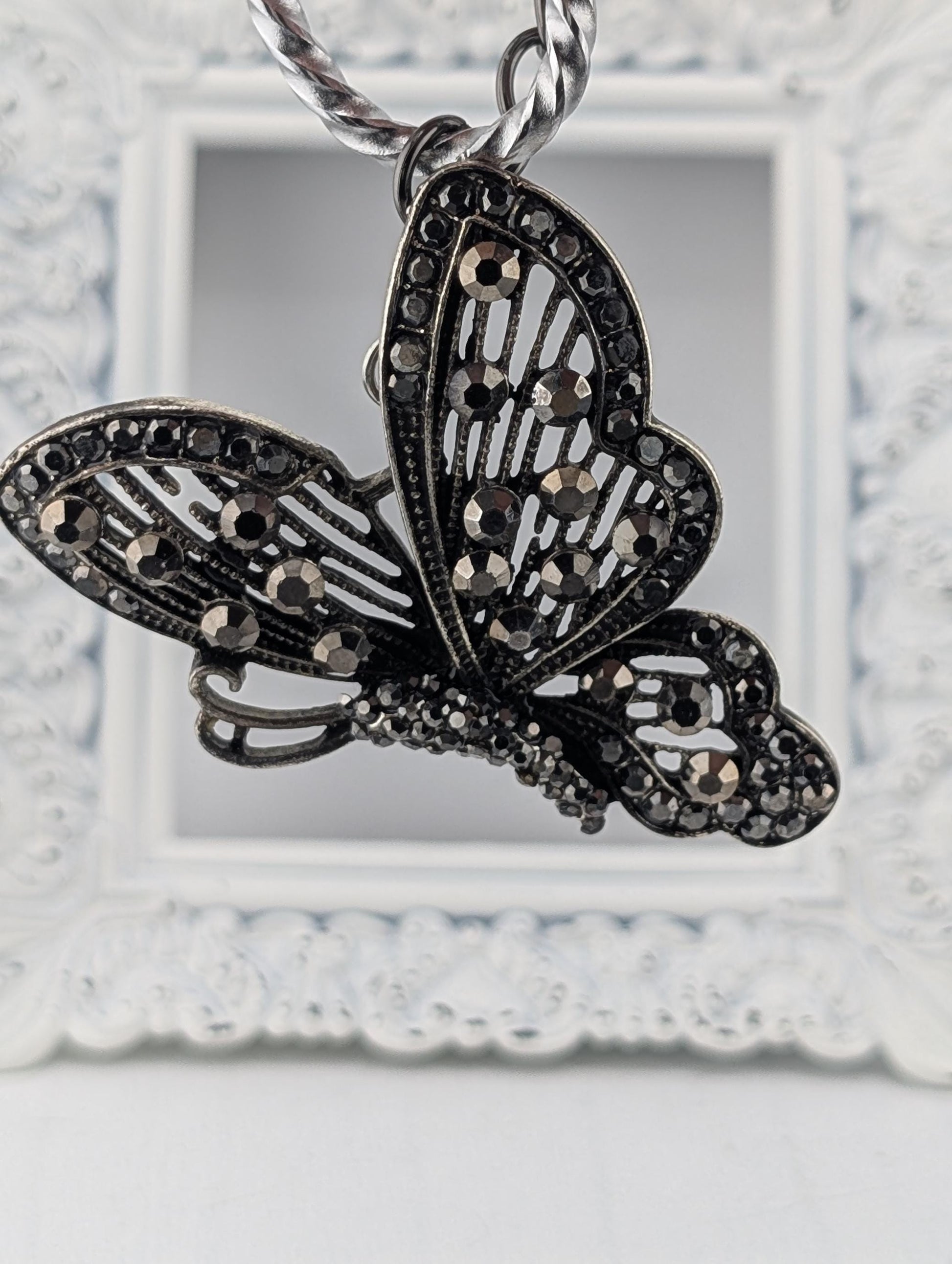 A decorative butterfly pendant with intricate black and silver designs, including rhinestones or crystals, hanging from a chain.