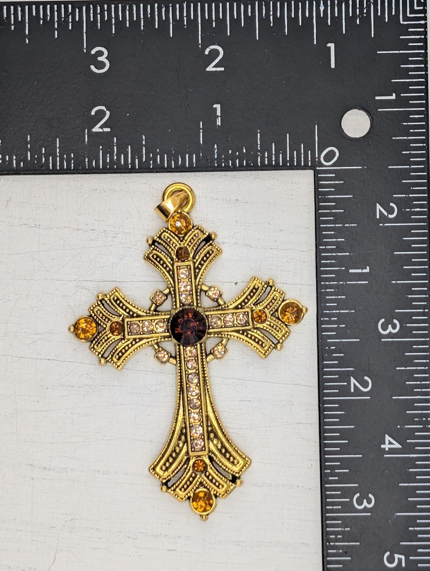 A gold cross pendant with gemstones is placed next to a ruler for size comparison.