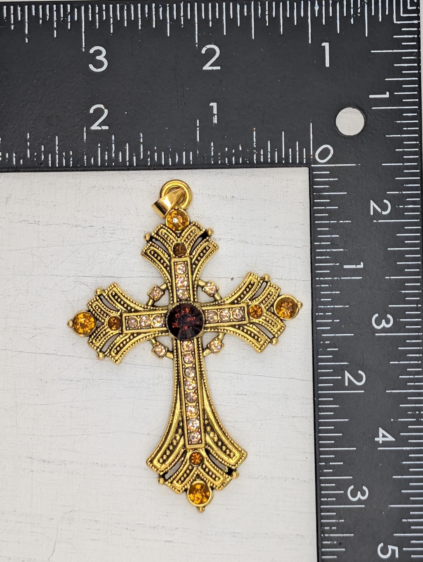 A gold cross pendant with gemstones is placed next to a ruler for size comparison.