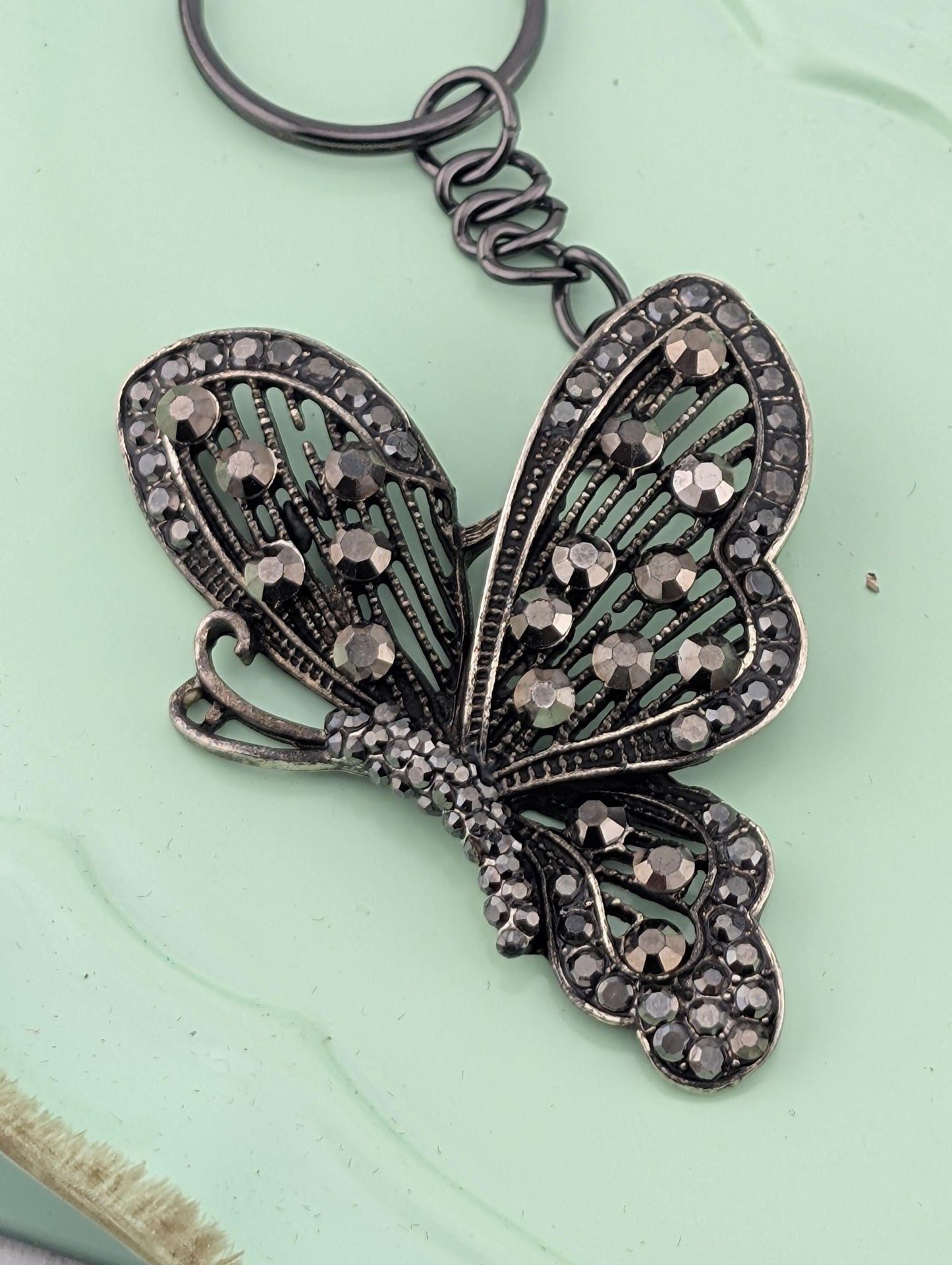 A decorative keychain in the shape of a butterfly, with intricate details and sparkling rhinestones or crystals adorning the wings and body.