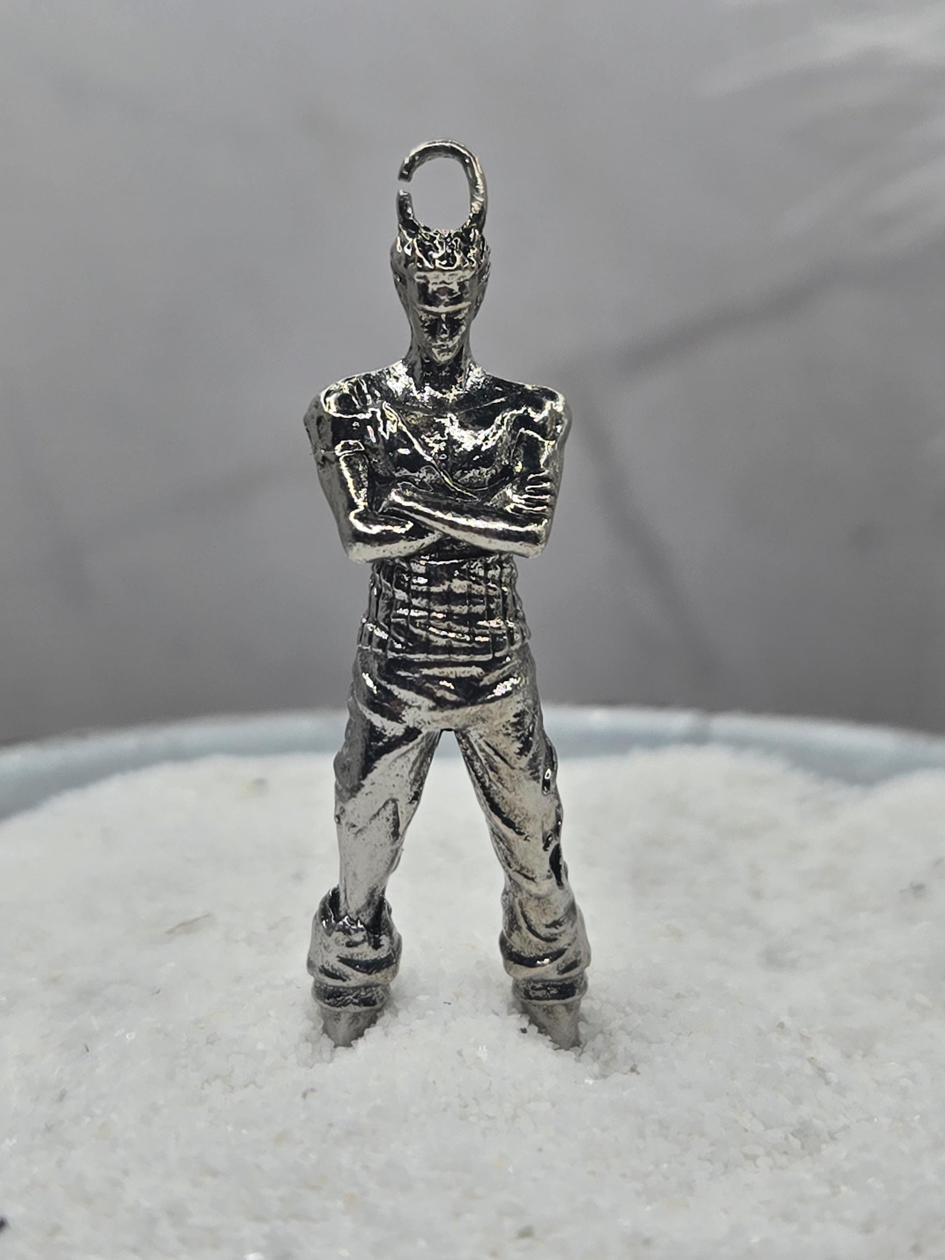 A silver-colored metal charm in the shape of a person, possibly a pendant or bracelet, is shown against a blurred background.