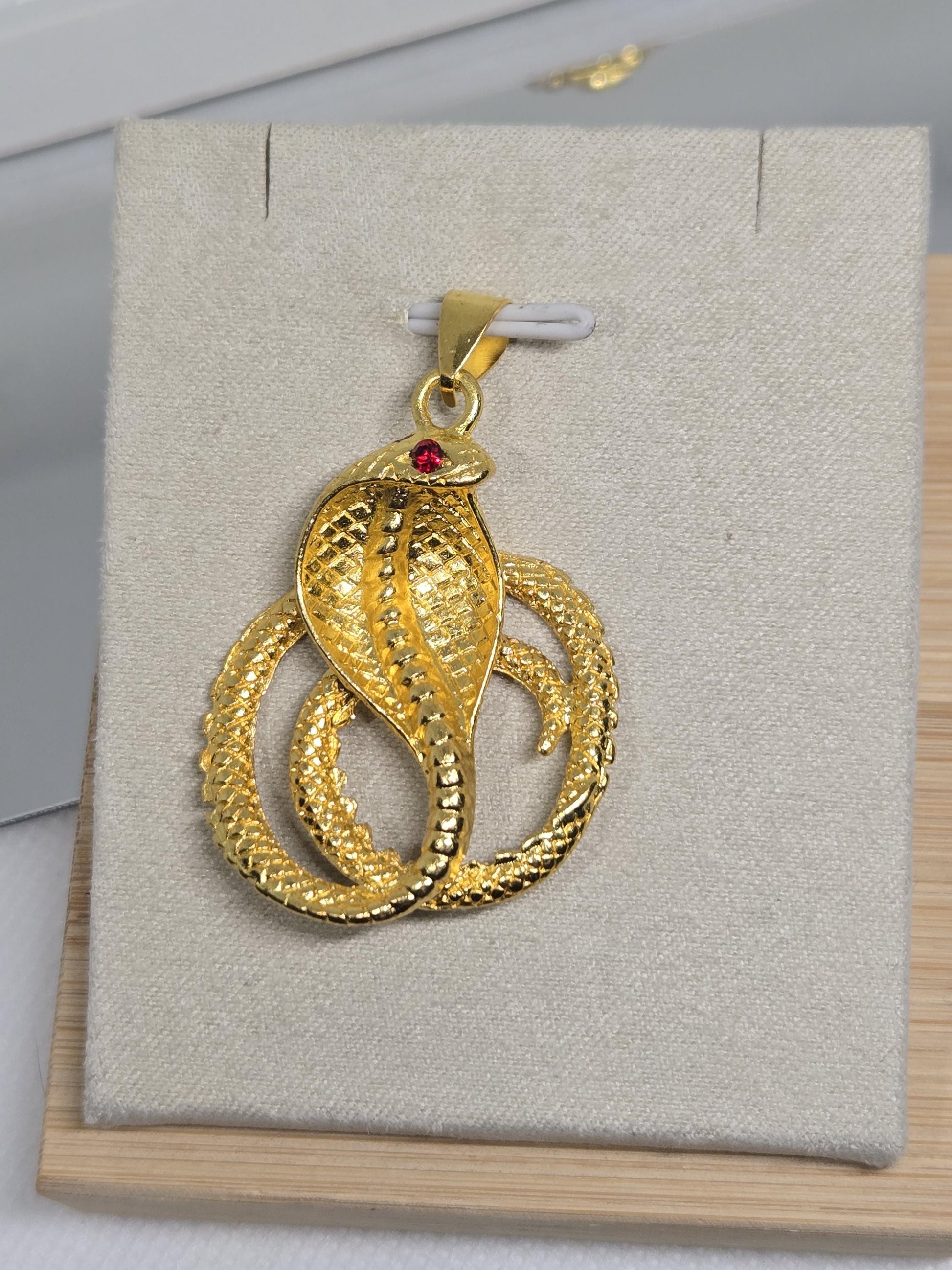 A gold snake pendant with a red gemstone is displayed on a white background.