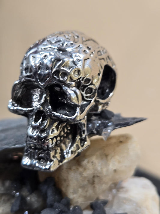 A close-up view of a silver-colored metal skull-shaped object, with a textured surface and a pointed top.