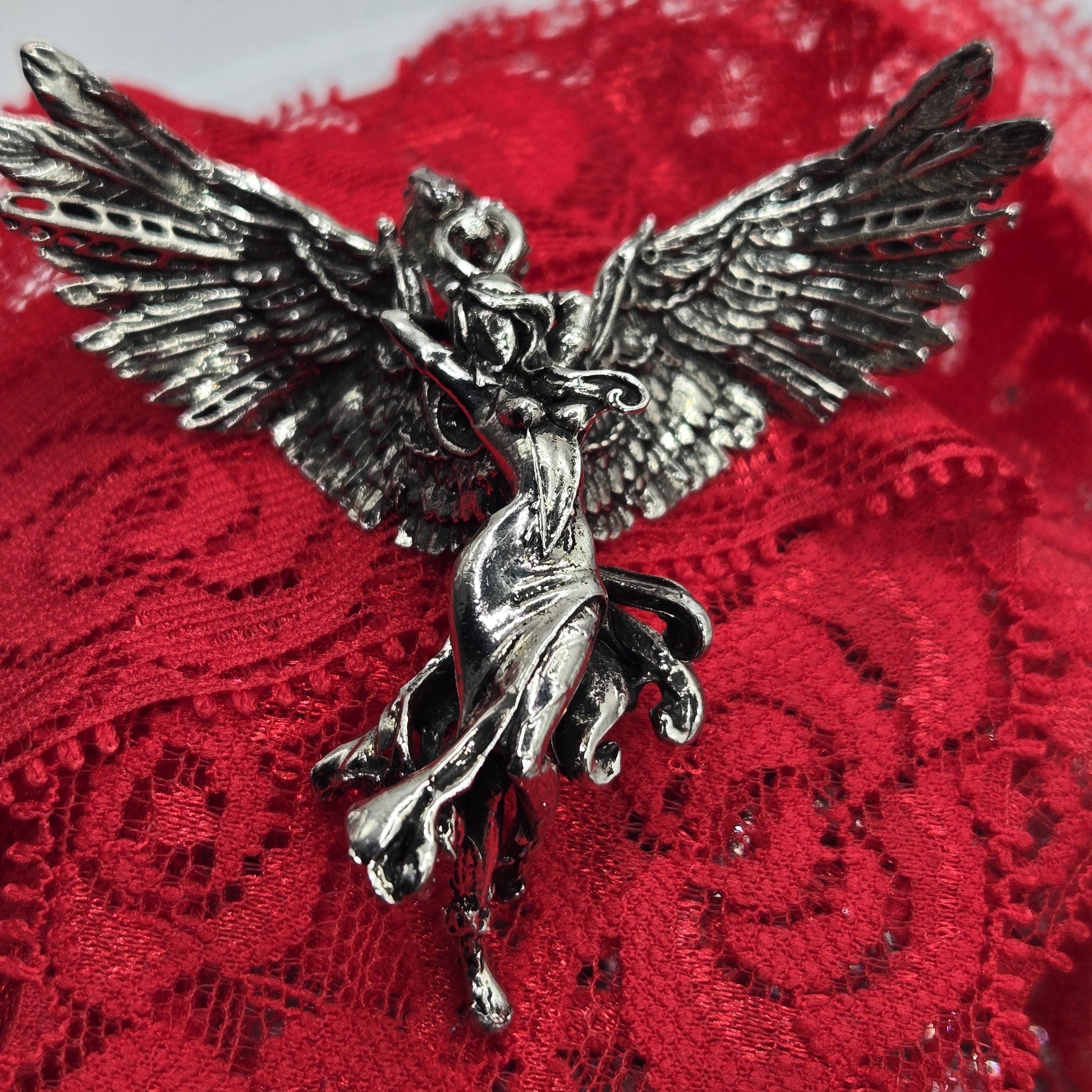 A close-up image of a silver metal pin or brooch with a winged design, resting on a red lace background.