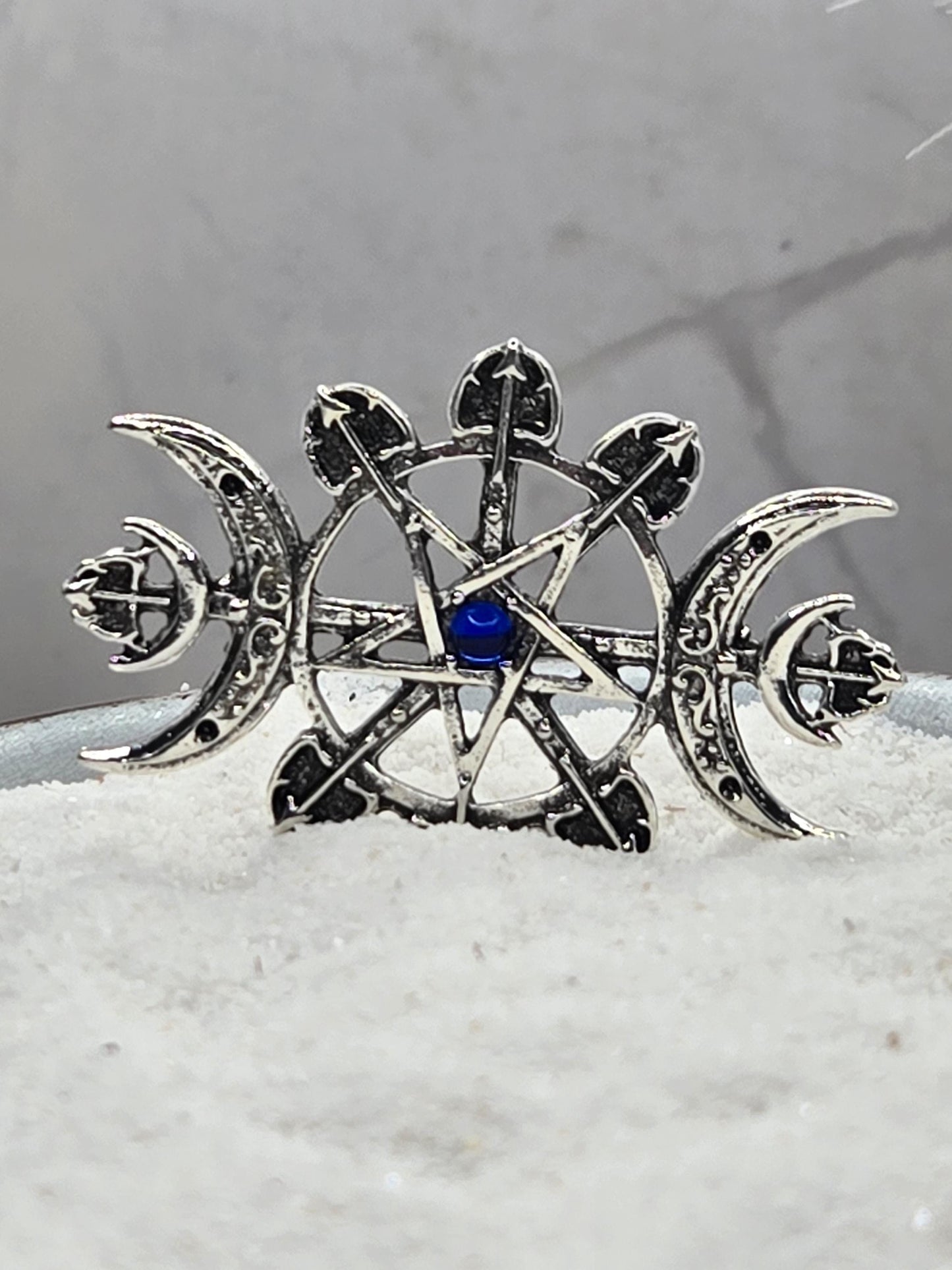 A silver metal pin with a blue gemstone, featuring intricate designs and symbols, such as a crescent moon and cross.