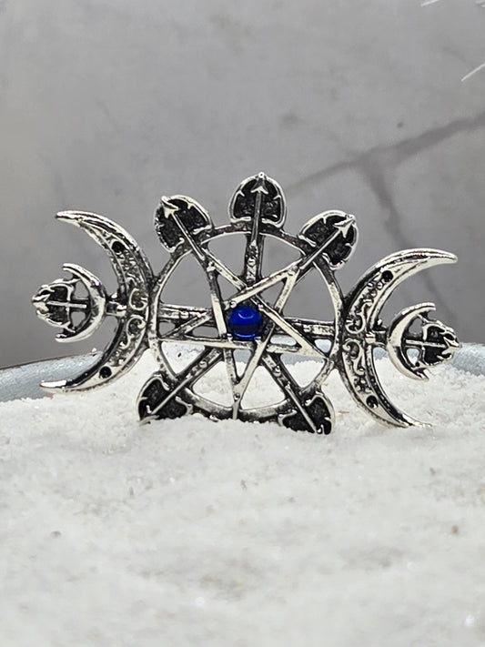 A silver metal pin with a blue gemstone, featuring intricate designs and symbols, such as a crescent moon and cross.
