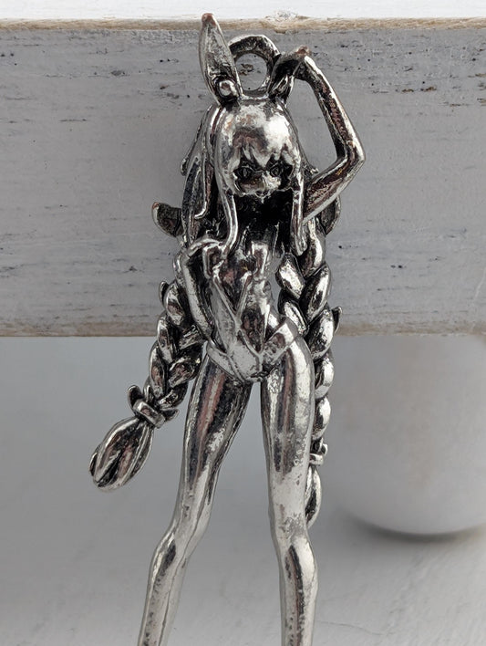 A silver-colored metal figurine of a woman with long braided hair, wearing a skirt and holding a broom.